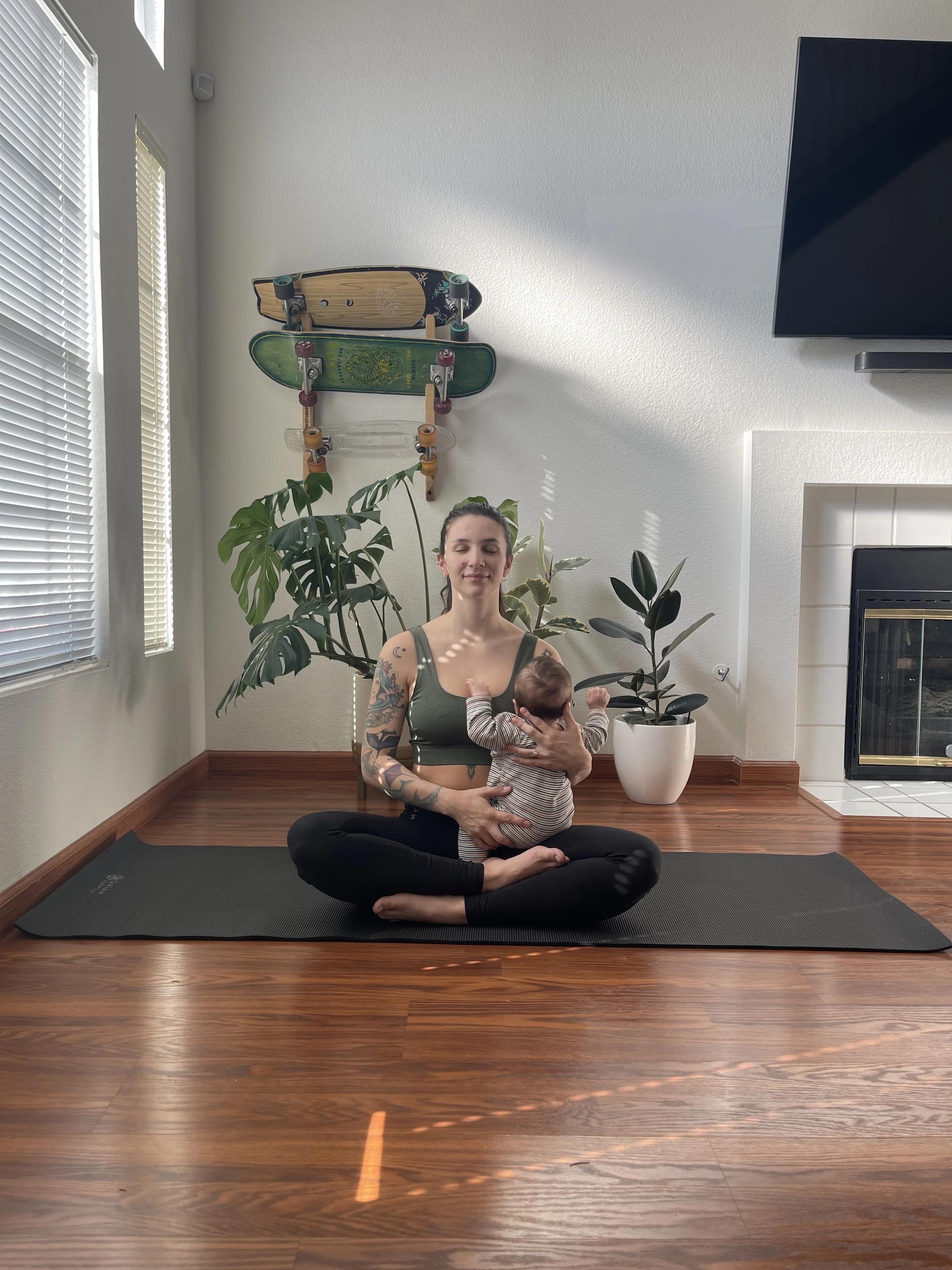 A woman practicing yoga in a living room with a baby. She is sitting cross-legged on a yoga mat, holding a baby sleeping on her chest. There are houseplants, skateboards mounted on the wall, a fireplace, and a television in the background.