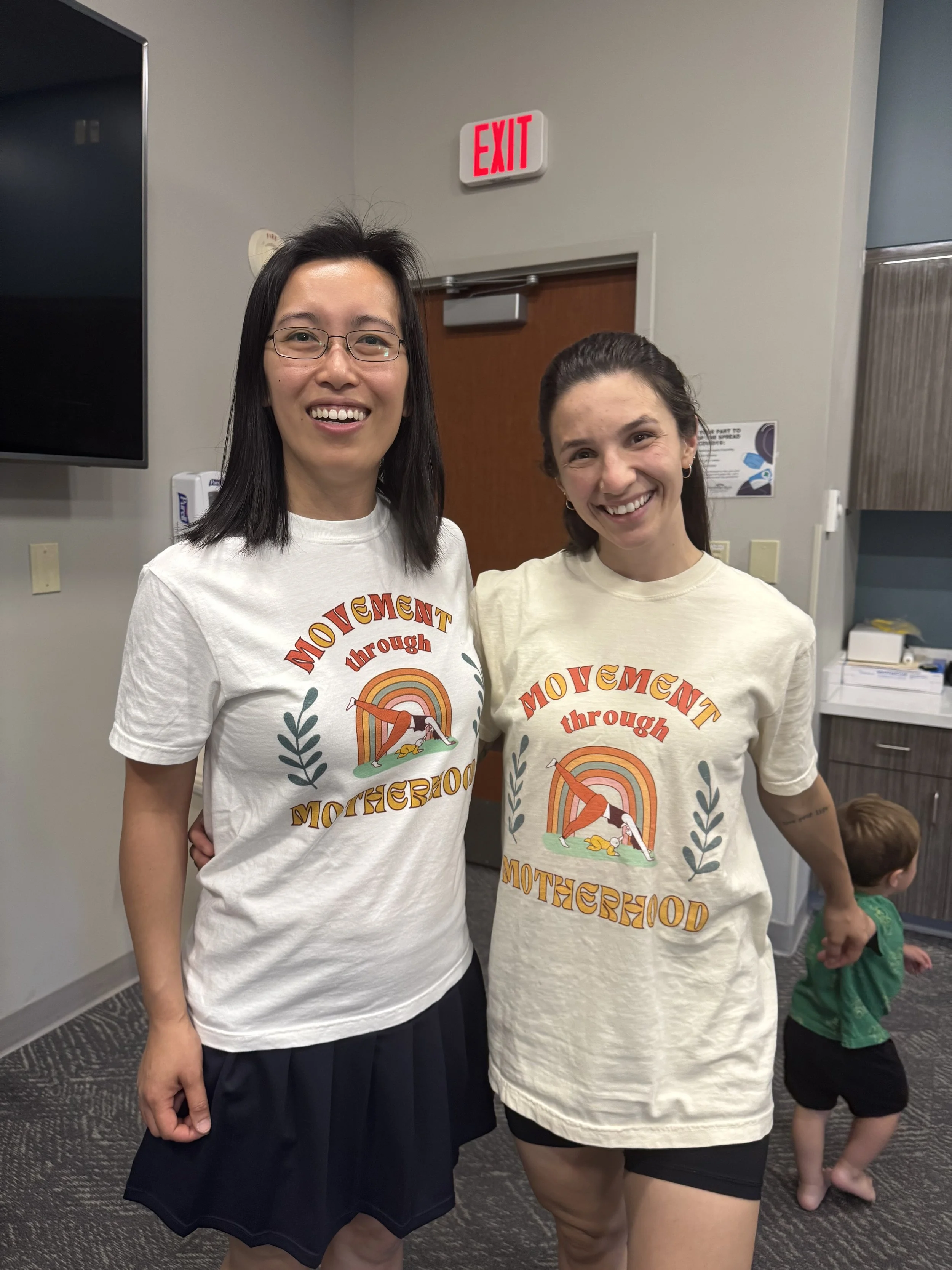 Two women wearing matching t-shirts that say "Movement through Motherhood" with a colorful graphic of a woman stretching, standing indoors, holding hands, with a young child in a green shirt and black shorts walking in the background.