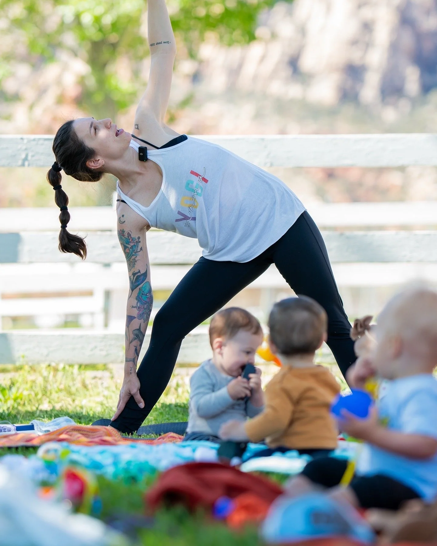 A woman practicing yoga outdoors with children sitting on a blanket nearby.