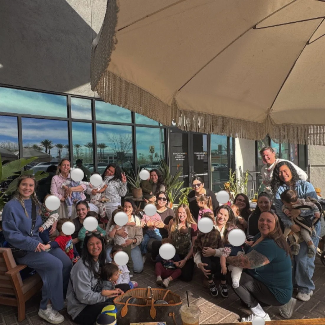 A group of people, including adults and children, gathered outside at a restaurant or café under a large beige umbrella with an outdoor patio. They are smiling and posing for a photo, with some holding children or making peace signs. There are plants, benches, and a glass storefront in the background.