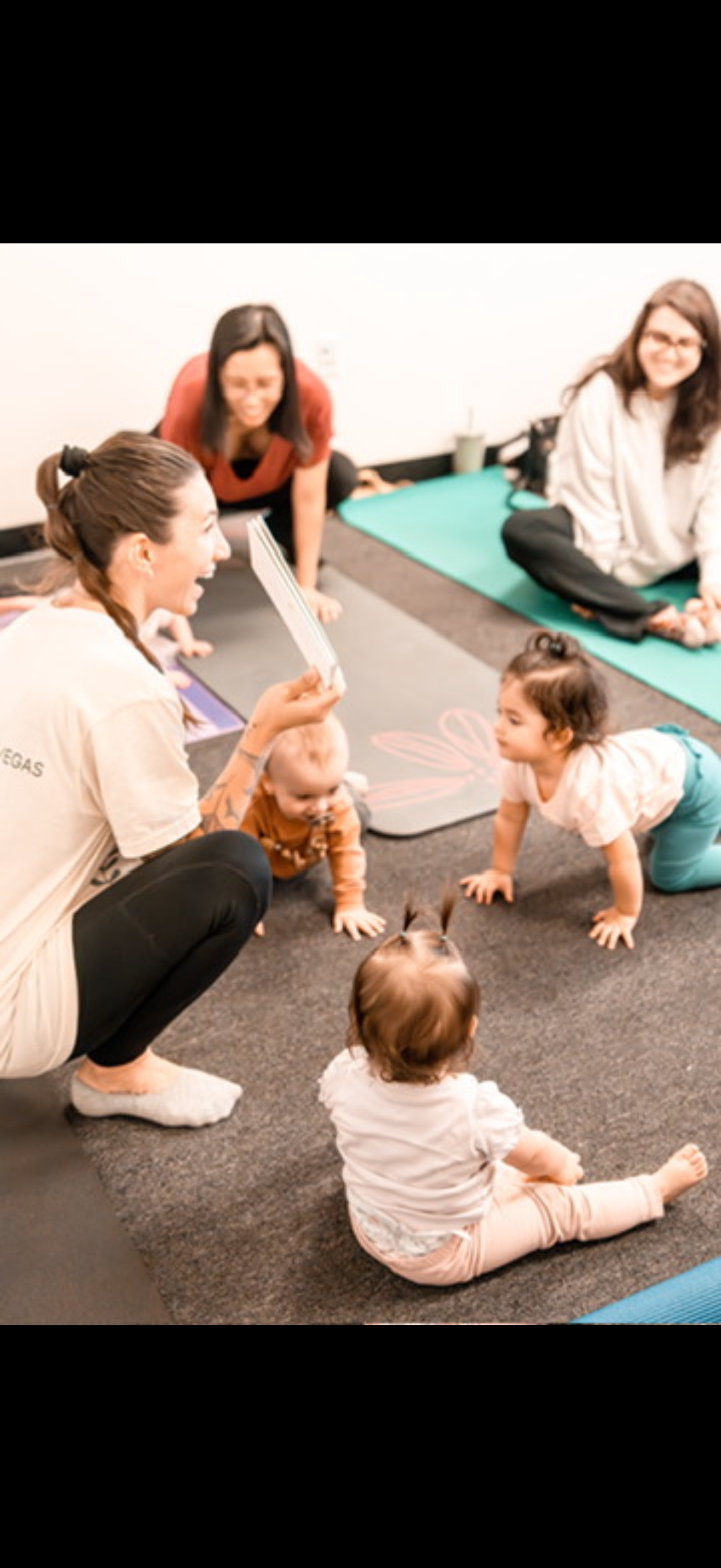 A group of women and children participating in a yoga or exercise class indoors, some on mats and others on the carpet, with one woman holding a yoga flashcard for kids.