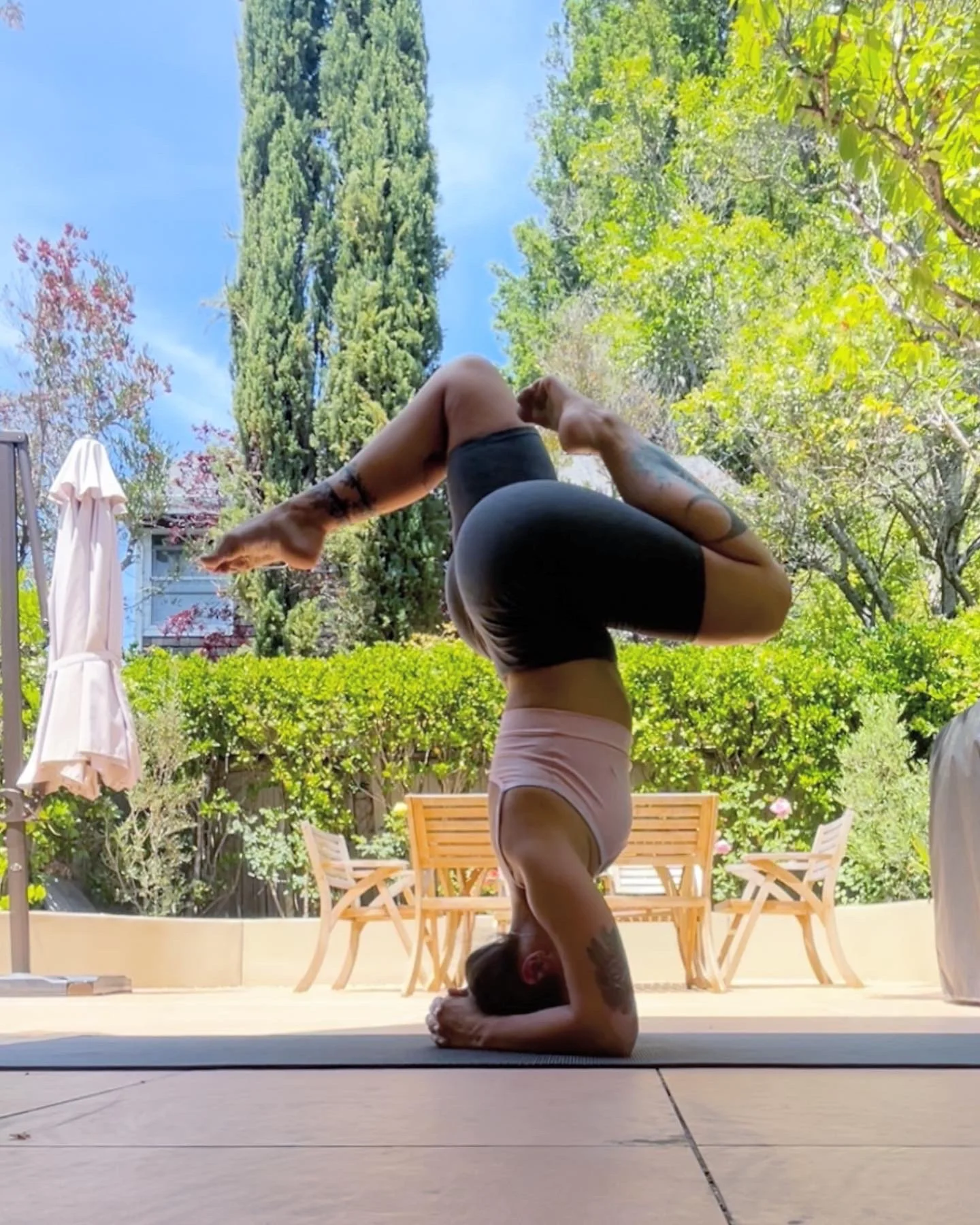 A woman practicing yoga outdoors in the headstand pose on a mat in a backyard with trees and outdoor furniture.