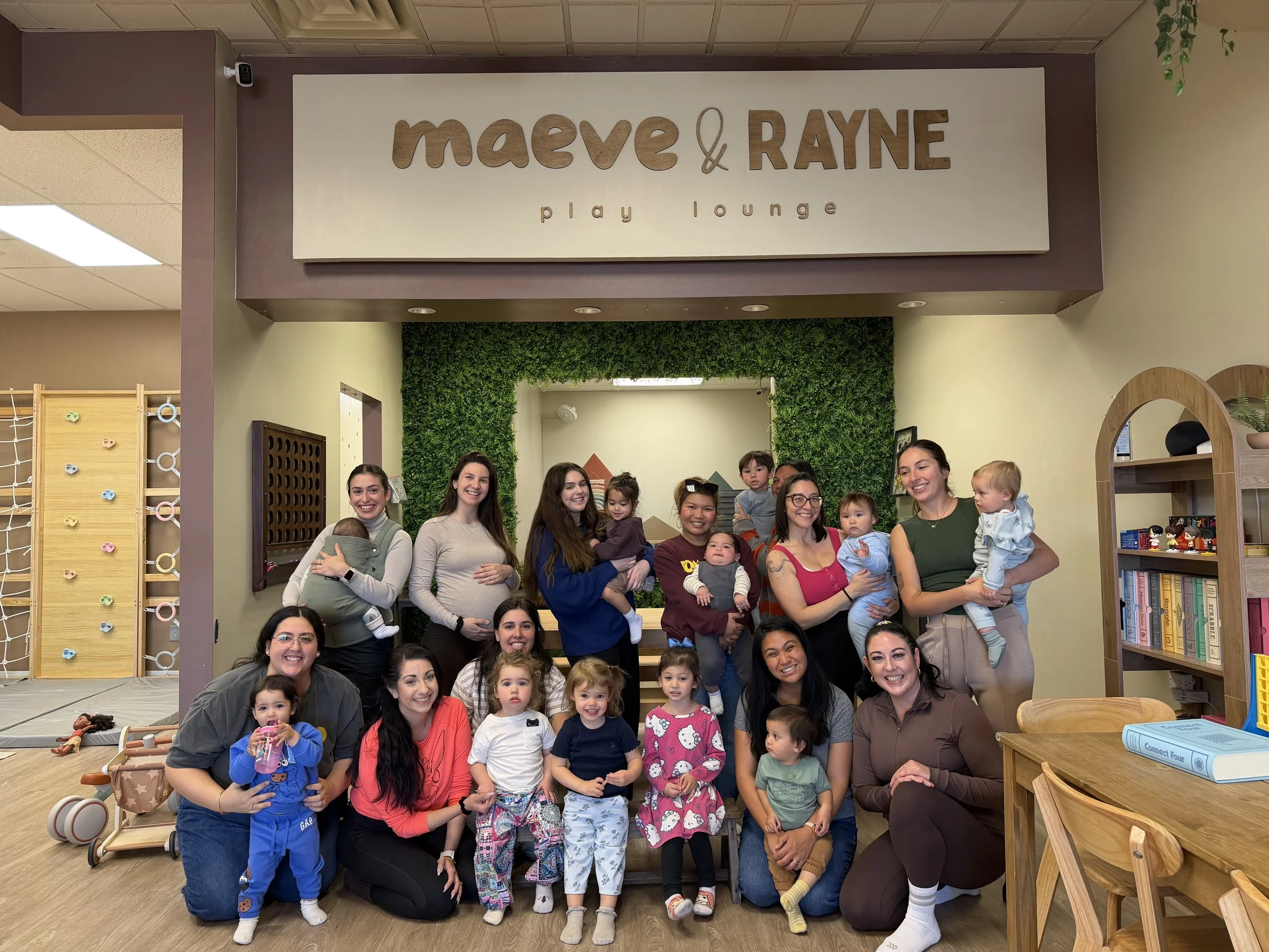 A group of women and children posing together inside a play lounge named 'Maeve & Rayne Play Lounge'. The women are holding babies and standing or sitting among a crowd of young children, with some smiling at the camera. The play lounge has a sign above that reads 'Maeve & Rayne', with a decorative green hedge in the background and wooden furniture on the sides.