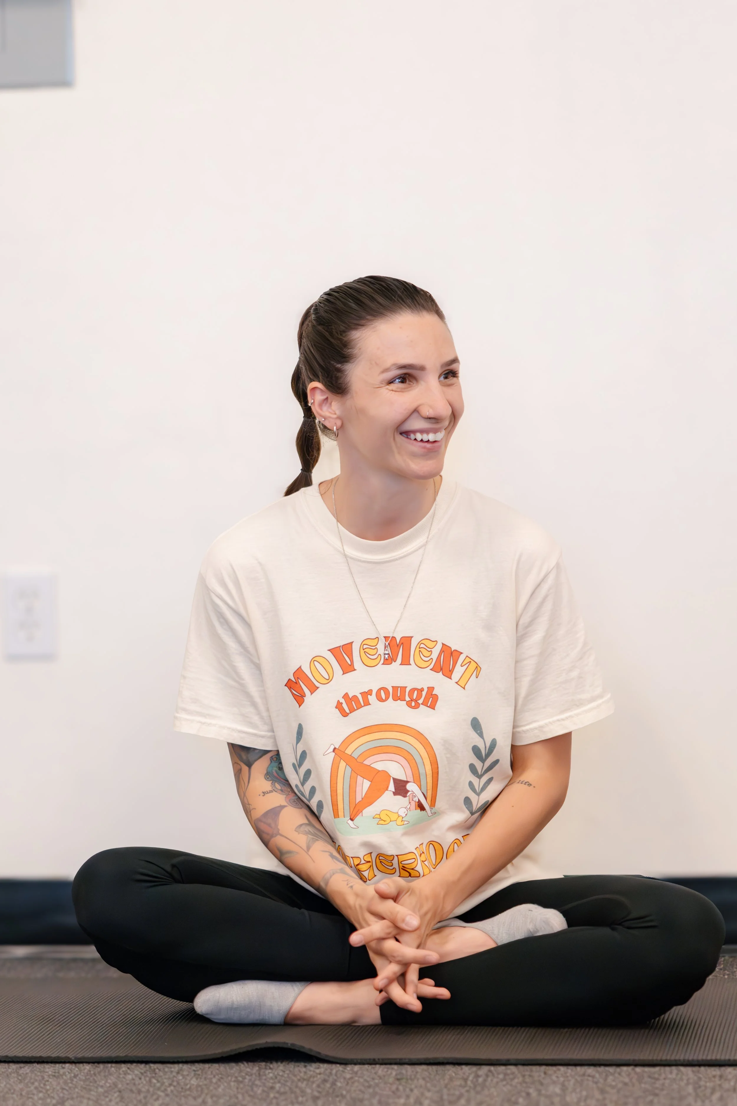 A woman with a ponytail sitting cross-legged on a black mat, smiling, wearing a beige T-shirt with the words 'Movement through Motherhood' and a graphic of a woman doing yoga, black leggings, and gray socks, in a room with a white wall.