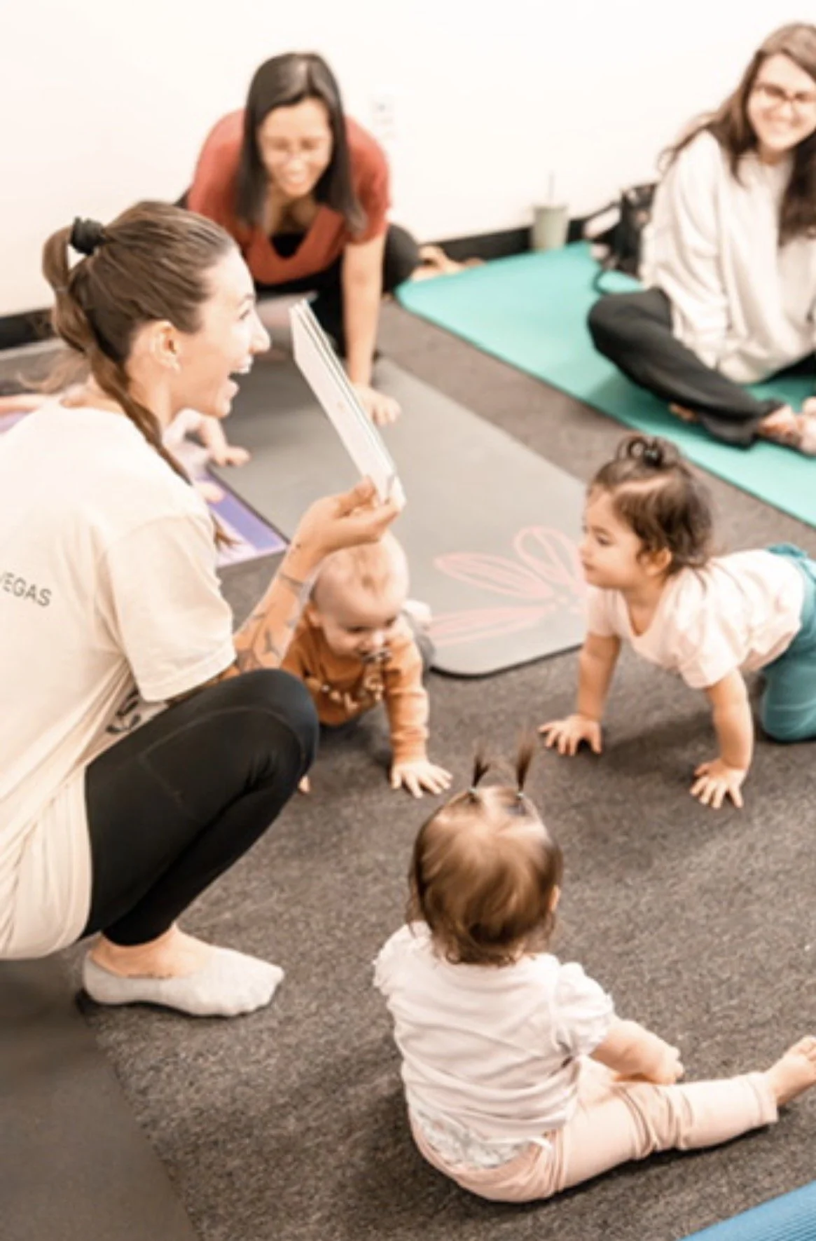 Group of women and children participating in a storytime or educational activity in a room with yoga mats and a carpeted floor.