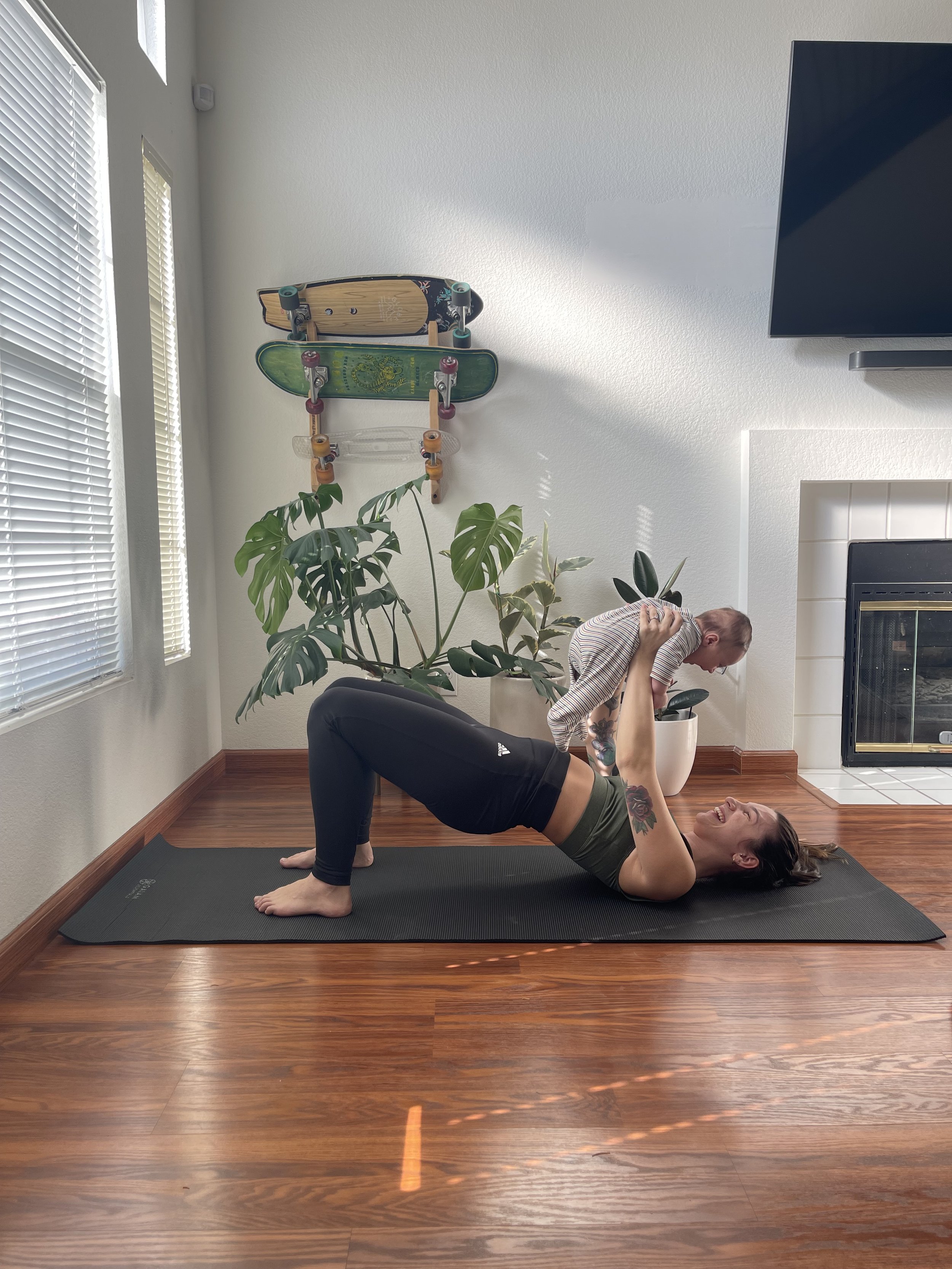 Woman doing a yoga pose on a yoga mat in a living room, holding a baby in the air, with a plant and skateboard decorations on the wall behind them.