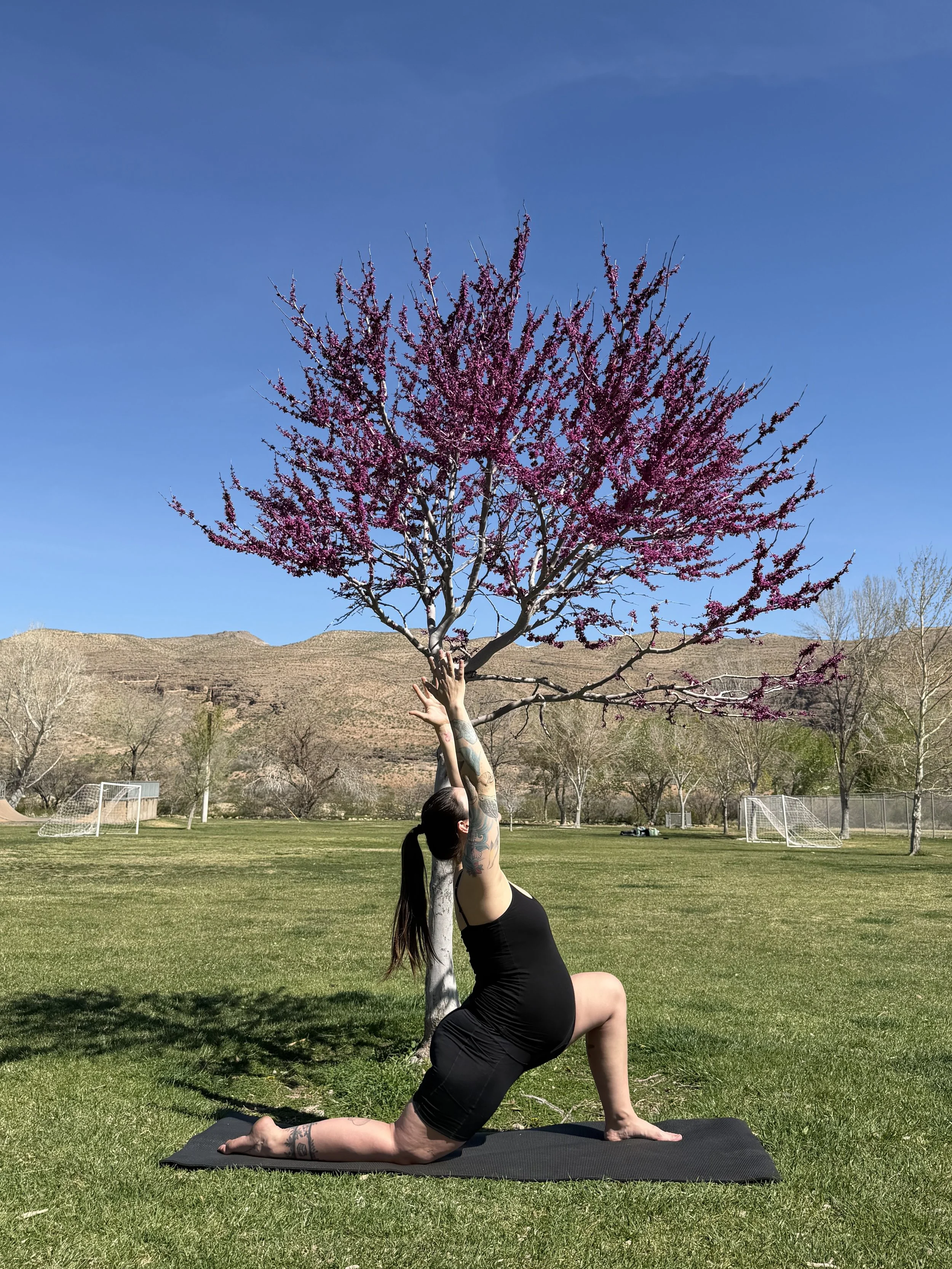 A woman practicing yoga outdoors on a black mat, doing a pose with one knee on the ground and arms extended upward, near a pink flowering tree in a park with a soccer field and hills in the background.