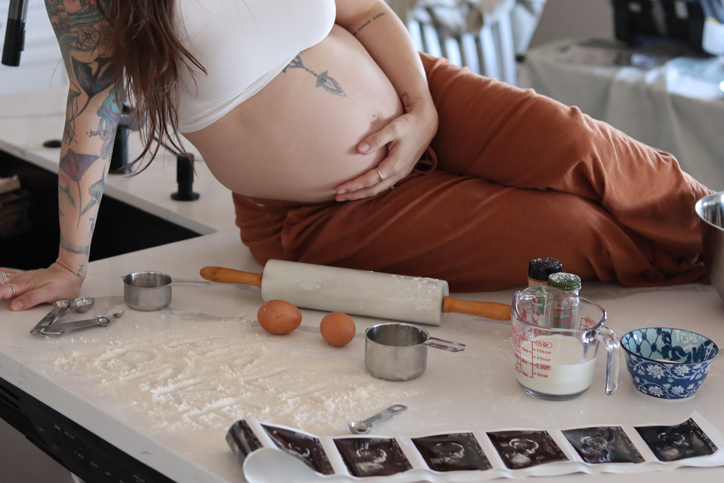 A pregnant woman with tattoos on her arm and side, leaning on a kitchen counter, with baking tools and ingredients, including eggs, rolling pin, measuring cups, and flour, in a home kitchen.