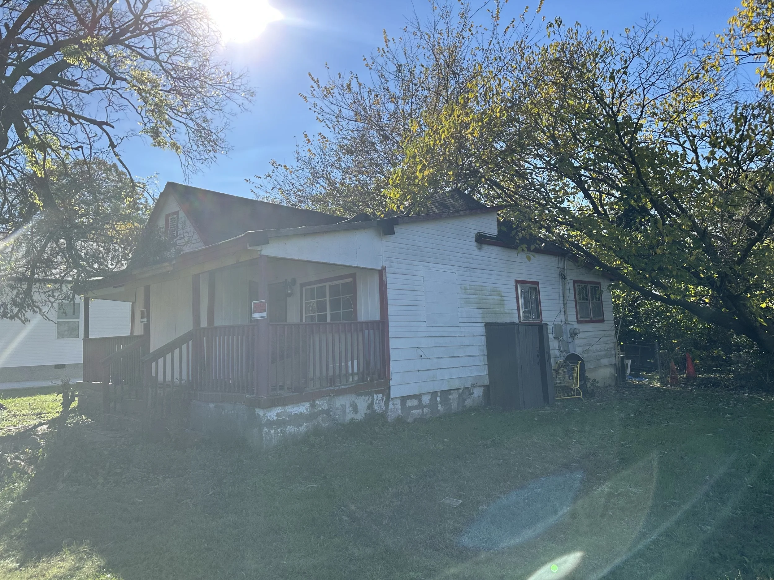 A small white house with a porch and red trim, surrounded by trees and grass, under a blue sky with the sun shining brightly.