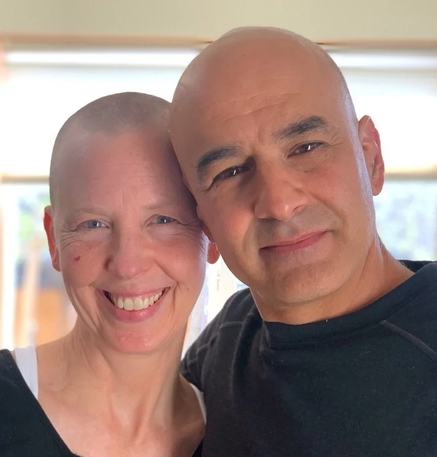 A smiling woman with a shaved head and a man with a shaved head and dark eyes, taking a close-up selfie together indoors.