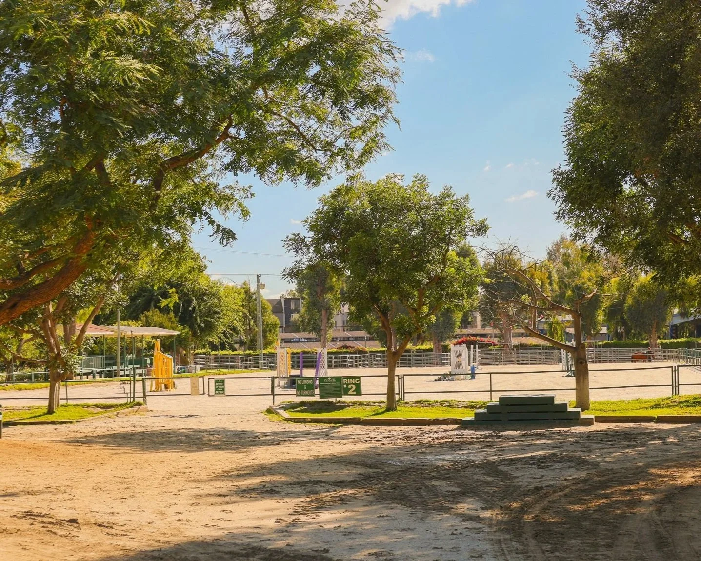 An horse riding arena with trees, a dirt path, and fenced play areas under a sunny sky.