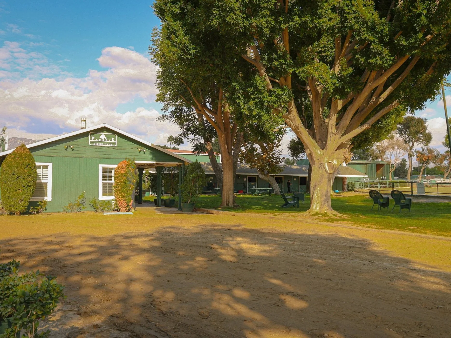 A park with a green building labeled 'Park' and outdoor seating, large trees providing shade, and a dirt pathway under a partly cloudy sky.