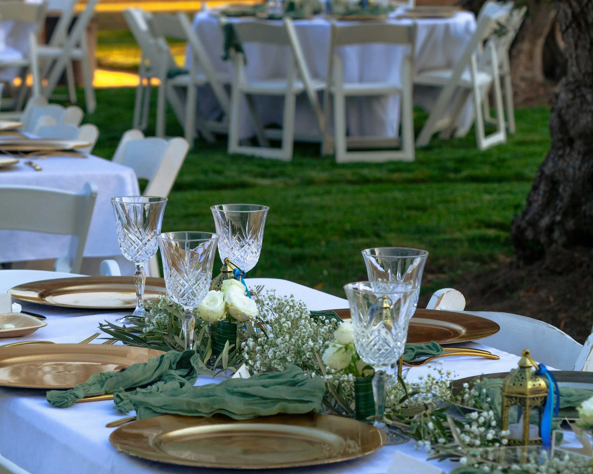 A decorated outdoor dining table with gold plates, crystal glasses, white flowers, and green napkins, set for an event in a grassy garden area with additional tables and chairs in the background.  Wedding venue, party venue, event center