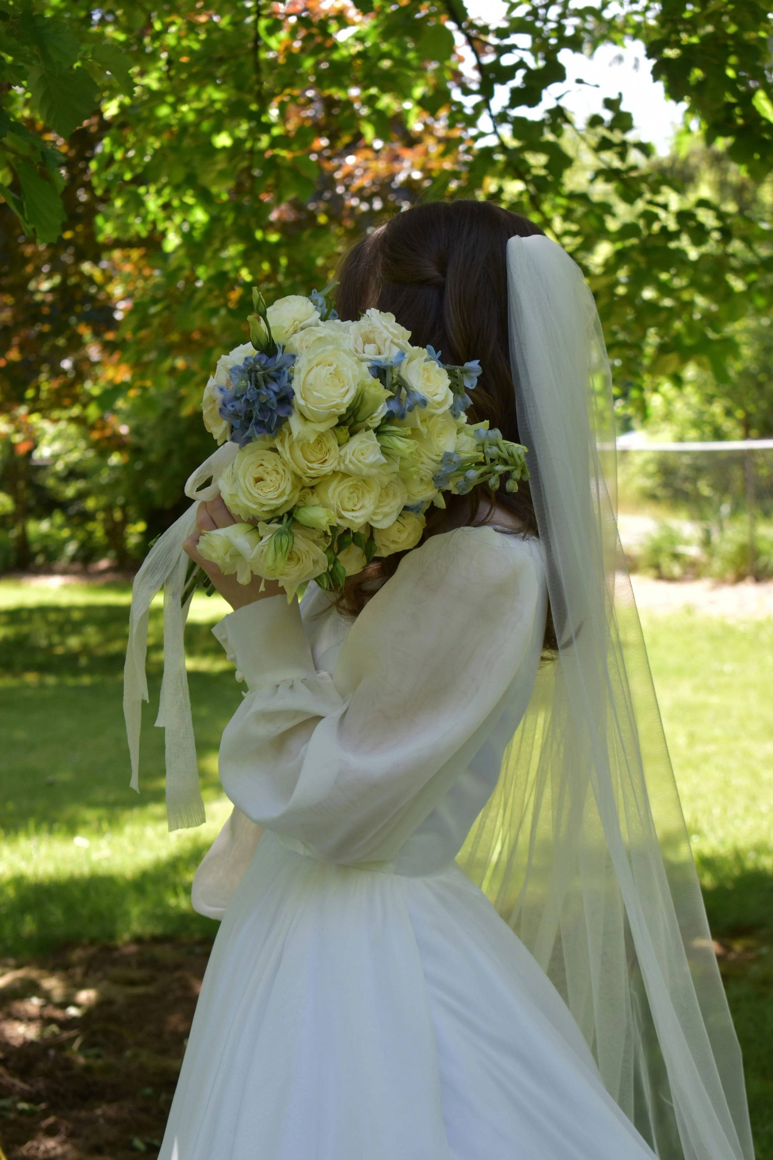 A bride in a white dress and veil holding a bouquet of white and blue flowers outdoors in a green park.