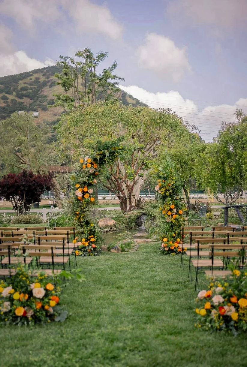 An outdoor wedding setup with a lush green lawn, wooden chairs, and a floral arch. The background features trees and a mountain landscape with cloudy sky.
