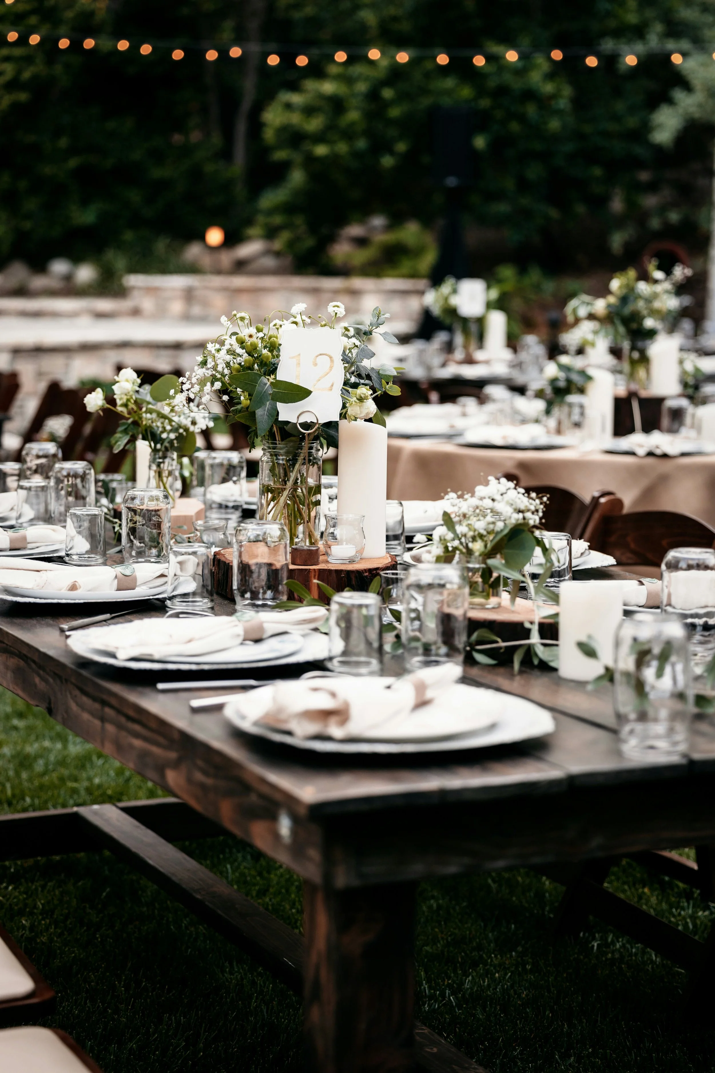 Outdoor wedding reception table decorated with white flowers, candles, and tableware, set in a garden or park.