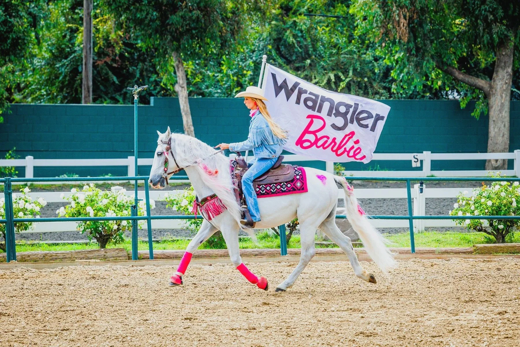 A young woman riding a white horse in an outdoor arena. She is wearing a cowboy hat, denim jacket, and jeans with pink accessories. The horse is decorated with a pink saddle blanket and pink leg wraps. Behind her, a large banner reads "Wrangler Barbie." Trees and a green fence surround the arena.