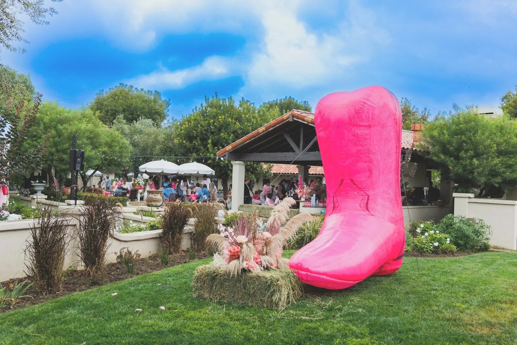 A large pink high heel shoe decoration in a garden at an outdoor gathering with people in the background, trees, and a pavilion with umbrellas.
