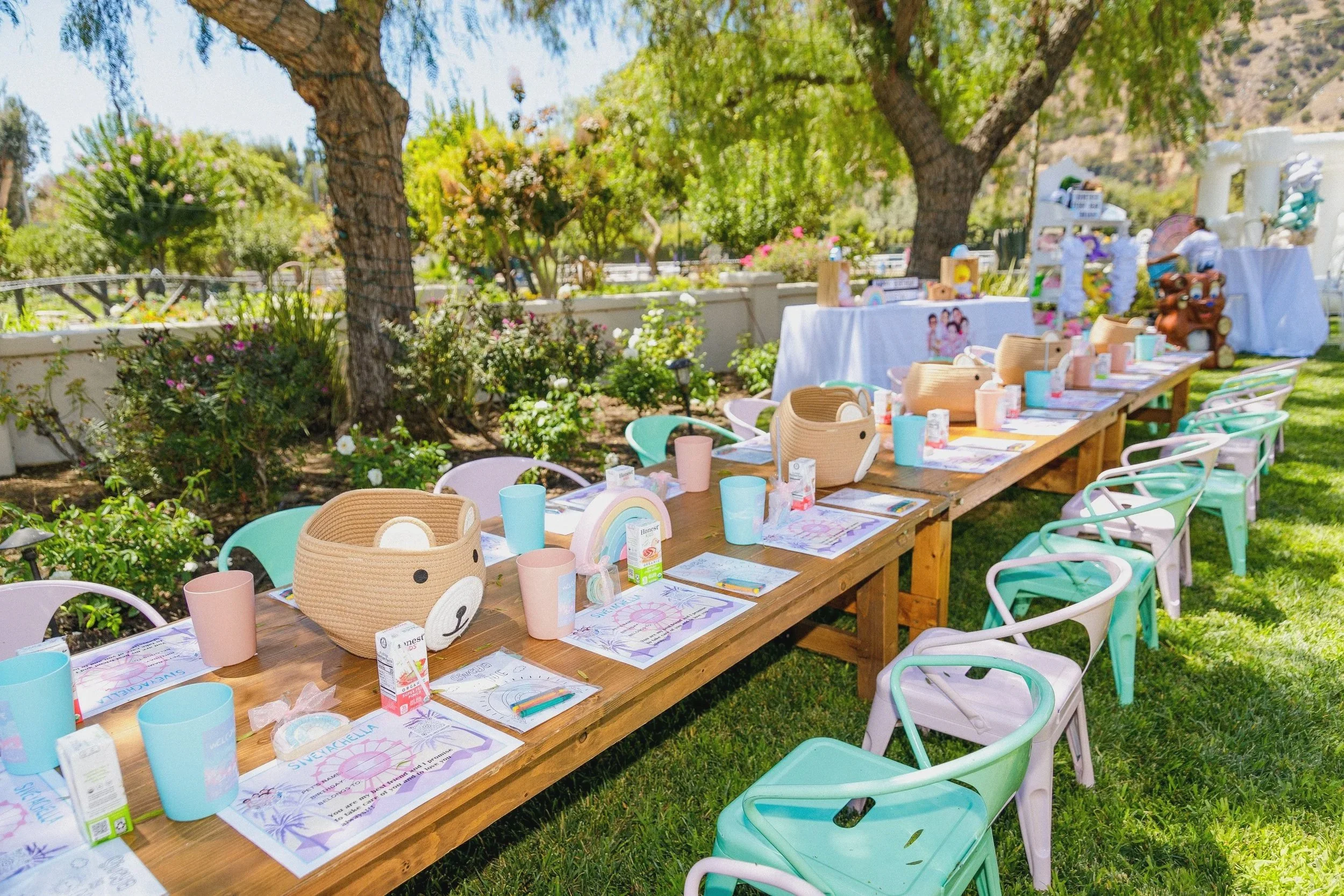 Decorated outdoor party table with pastel-colored chairs, placemats, cups, and themed decorations in a garden setting.
