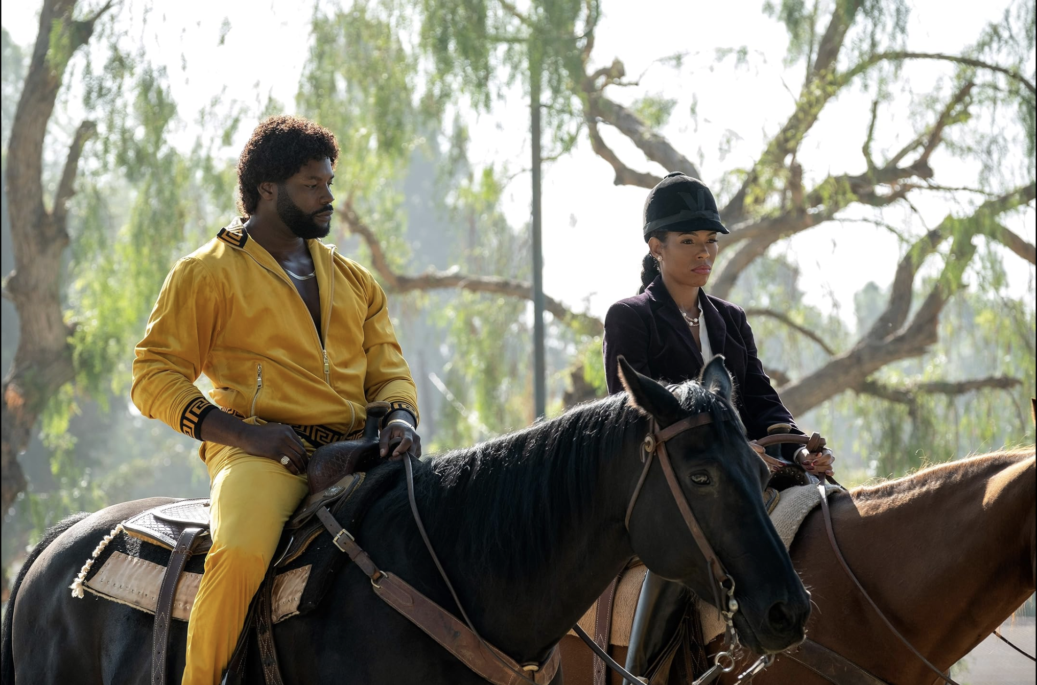 Man and woman riding horses outdoors, man in yellow jacket and woman in dark jacket and black cap, trees in background scene from a tv show at a horse barn, multi film location and photo shoot
