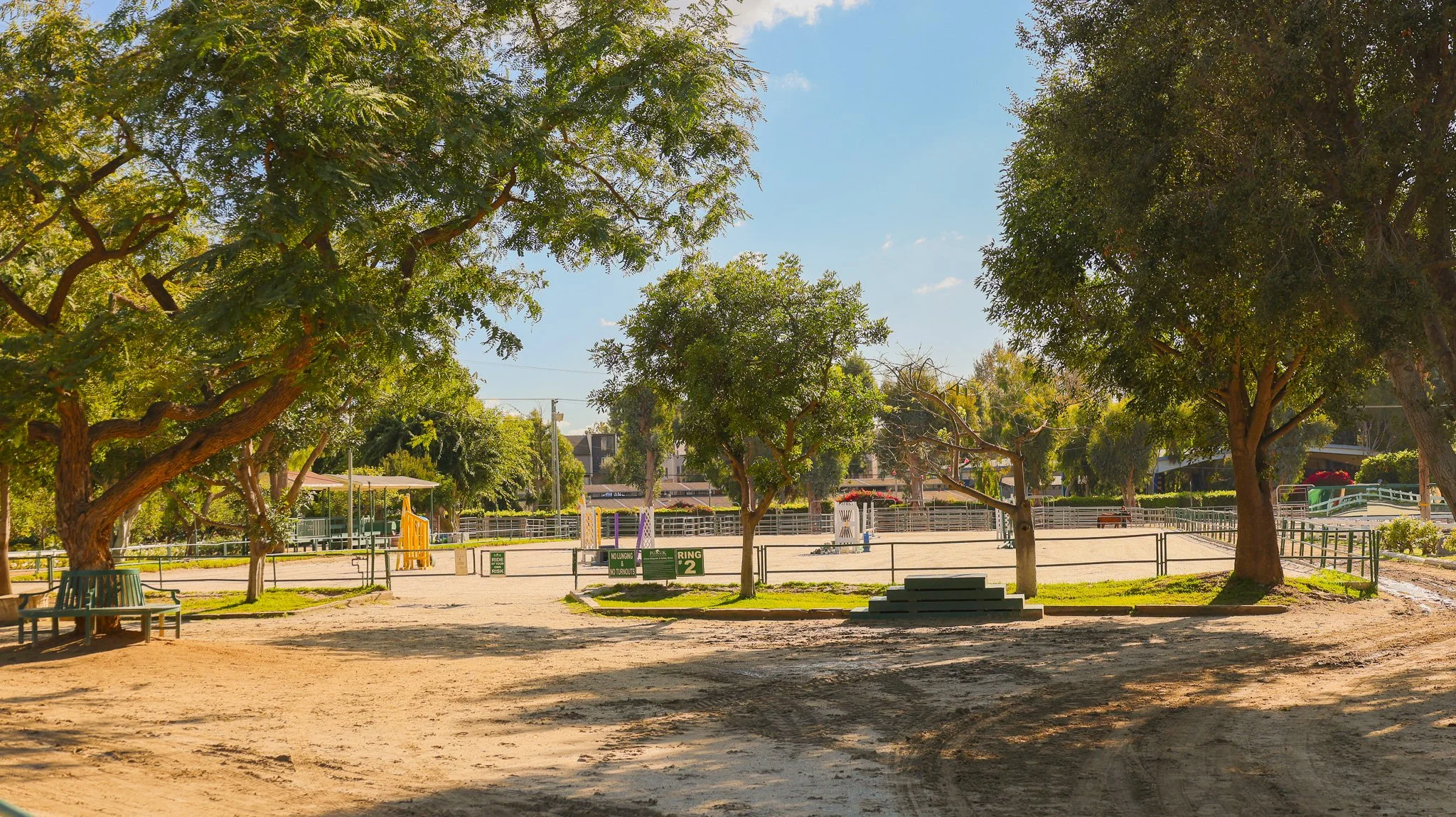 An outdoor playground area surrounded by trees, with equestrian show jumps, benches, and signs, under a clear blue sky.