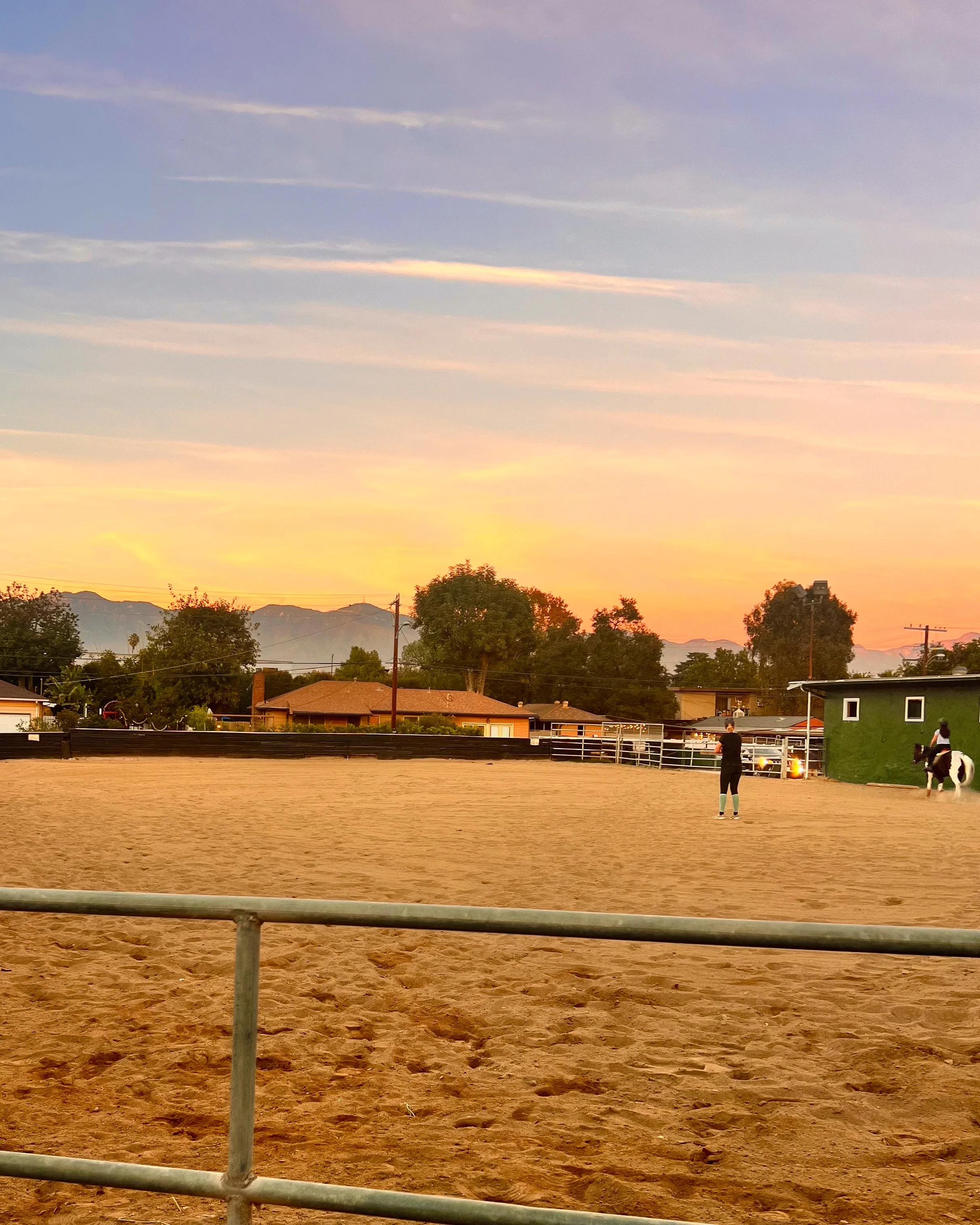 An outdoor riding arena during sunset with two people, one on horseback and one standing nearby, surrounded by a safety fence, with trees, houses, and mountains in the background.
