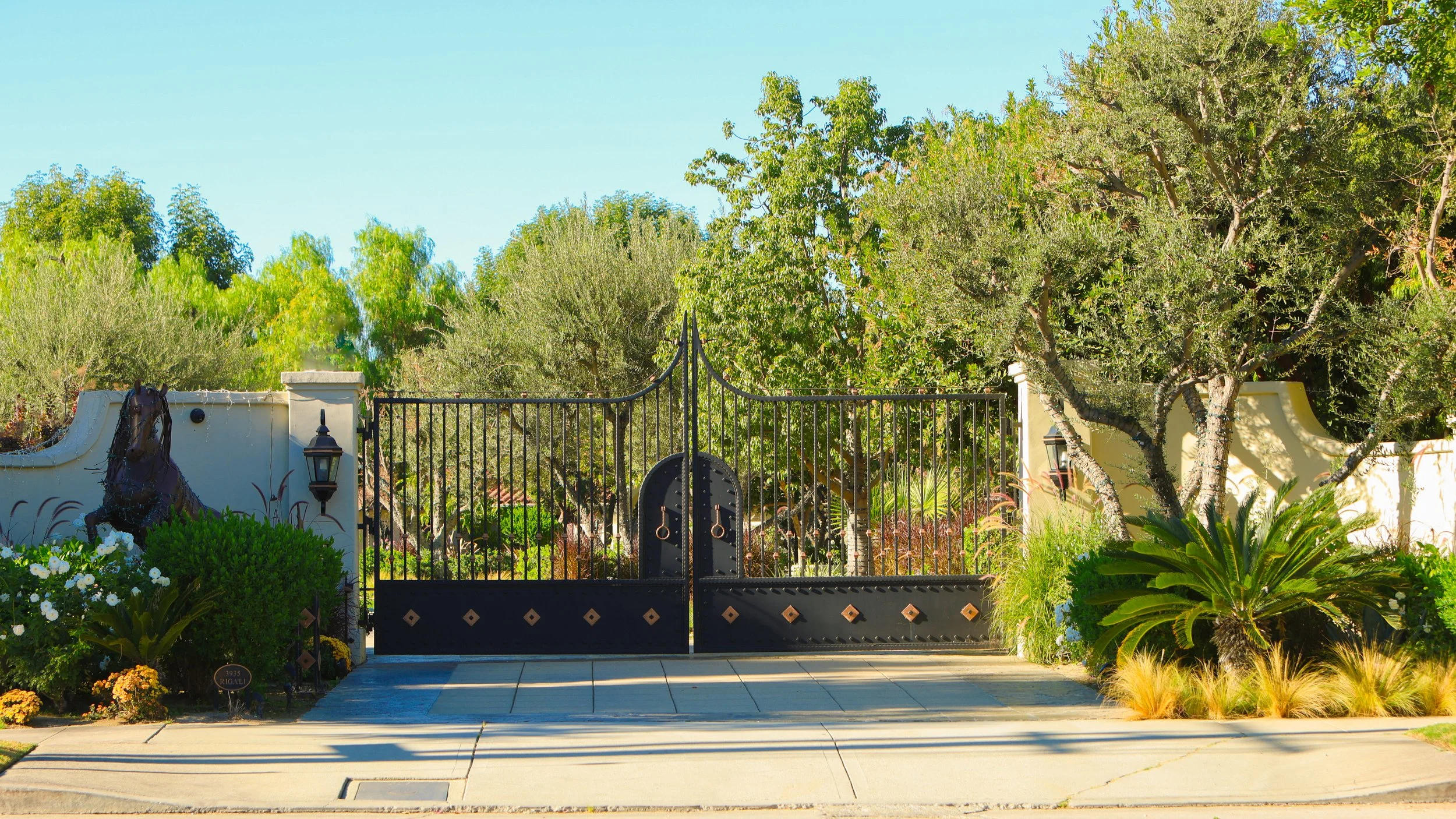 Black wrought-iron gate with decorative diamond patterns and two rings at the top, flanked by white curved wall sections with lanterns, surrounded by lush trees and plants, including large leafy bushes and a sago palm, under a clear blue sky.