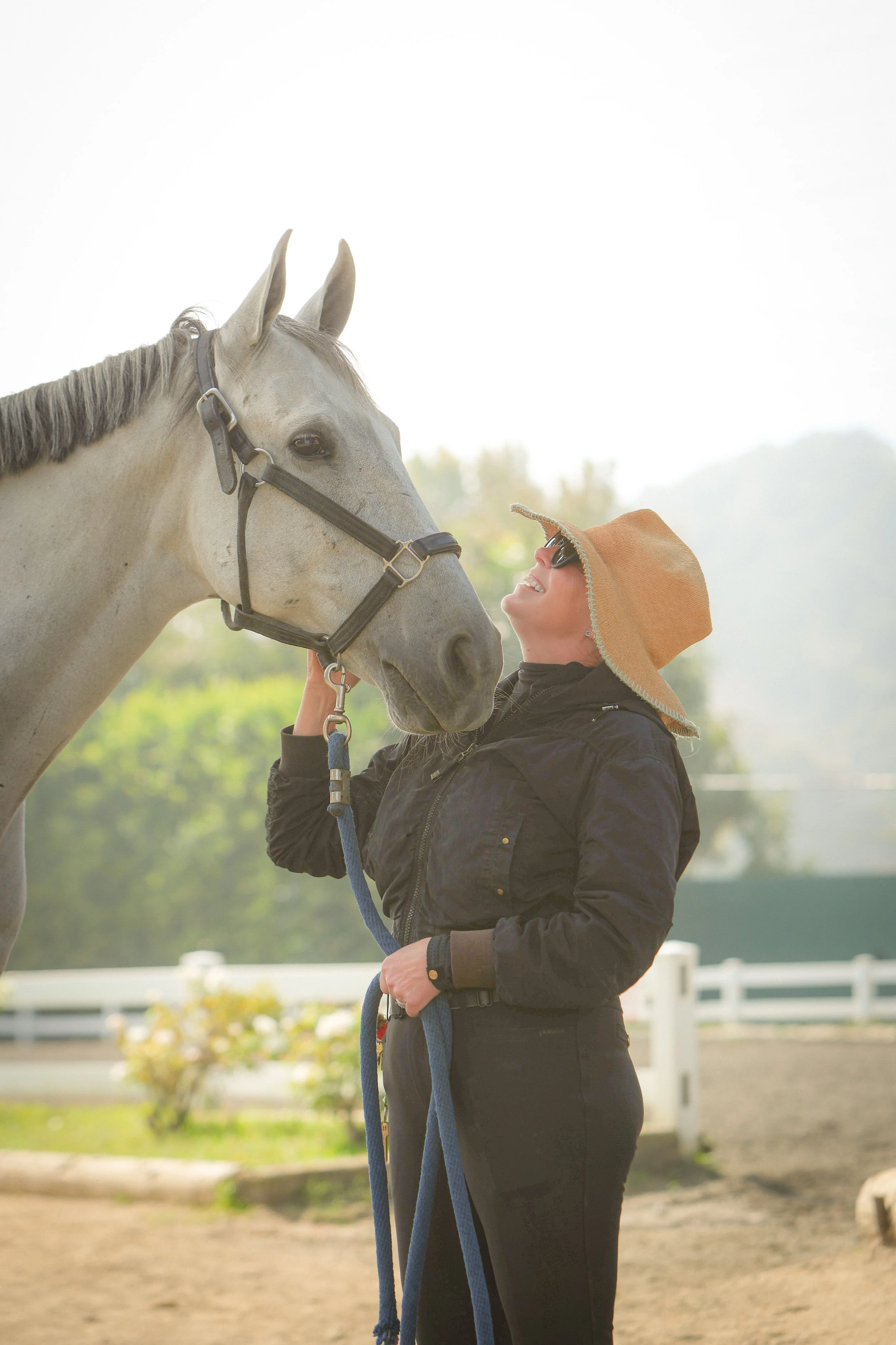 A woman in a black jacket and wide-brimmed hat smiling at a white horse at a riding area.