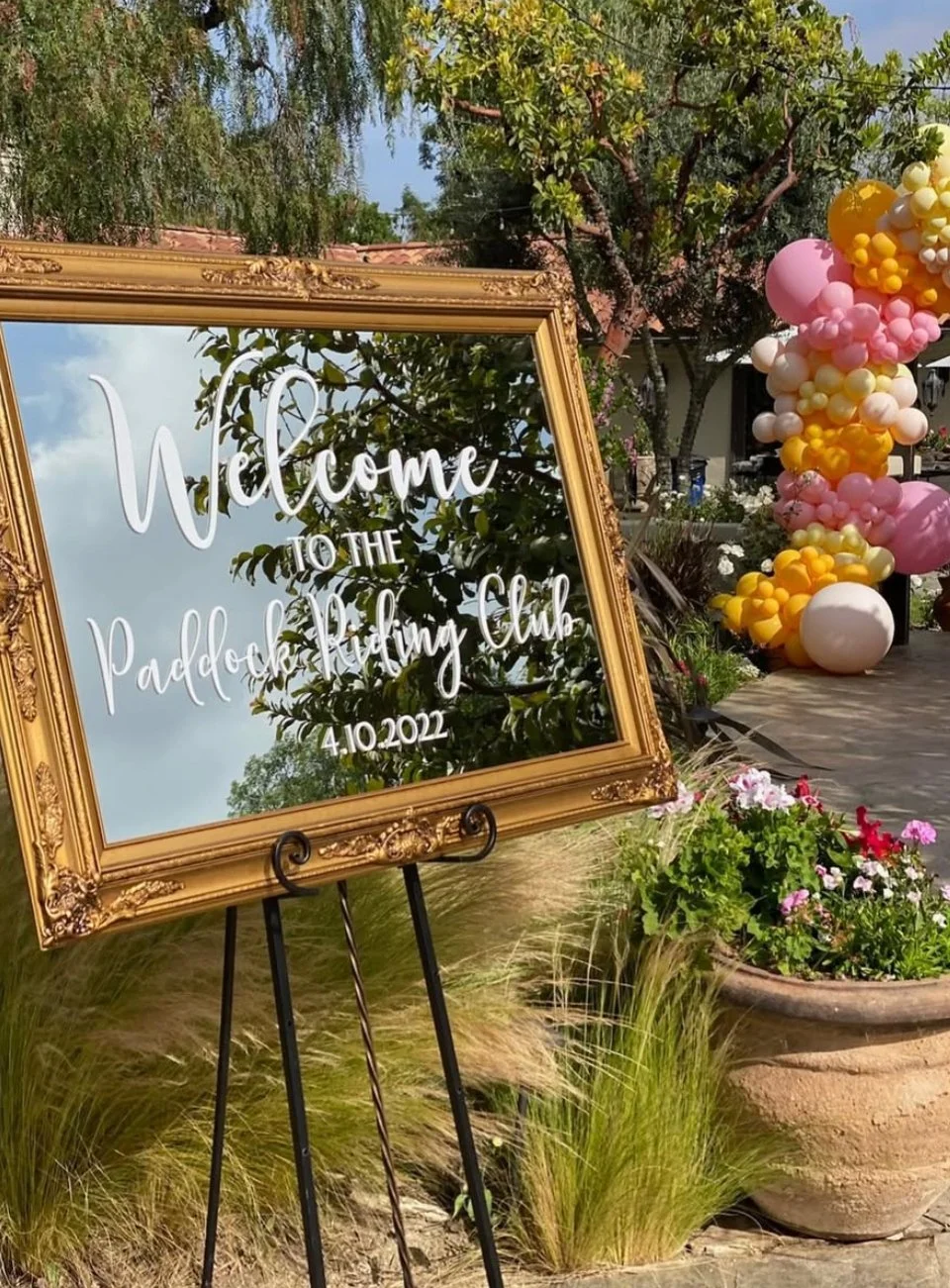 Decorative outdoor setting featuring a gold framed mirror stating 'Welcome to the Paddock Riding Club, 4.10.2022,' with colorful balloons and potted flowers in the background. Perfect for a wedding venue or Bat Mitzvah or Baby shower