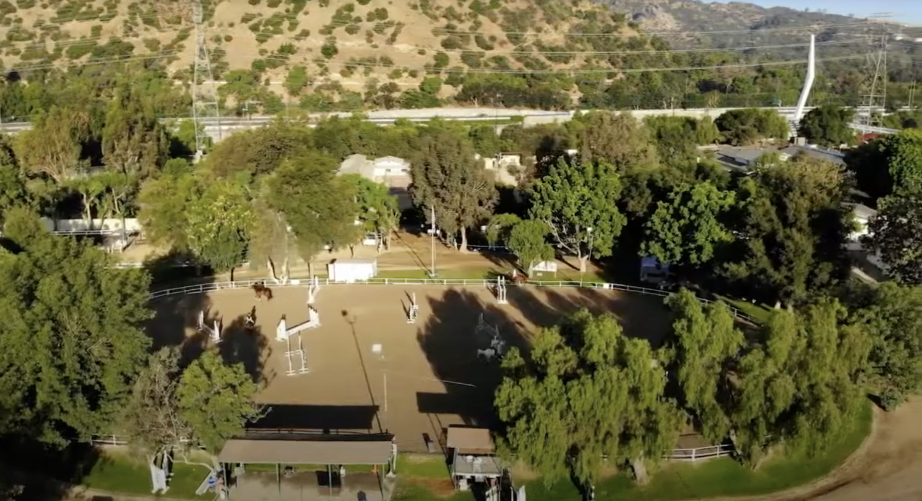 Aerial view of a horse riding arena surrounded by trees with a fenced perimeter, nearby buildings, and a hillside in the background.
