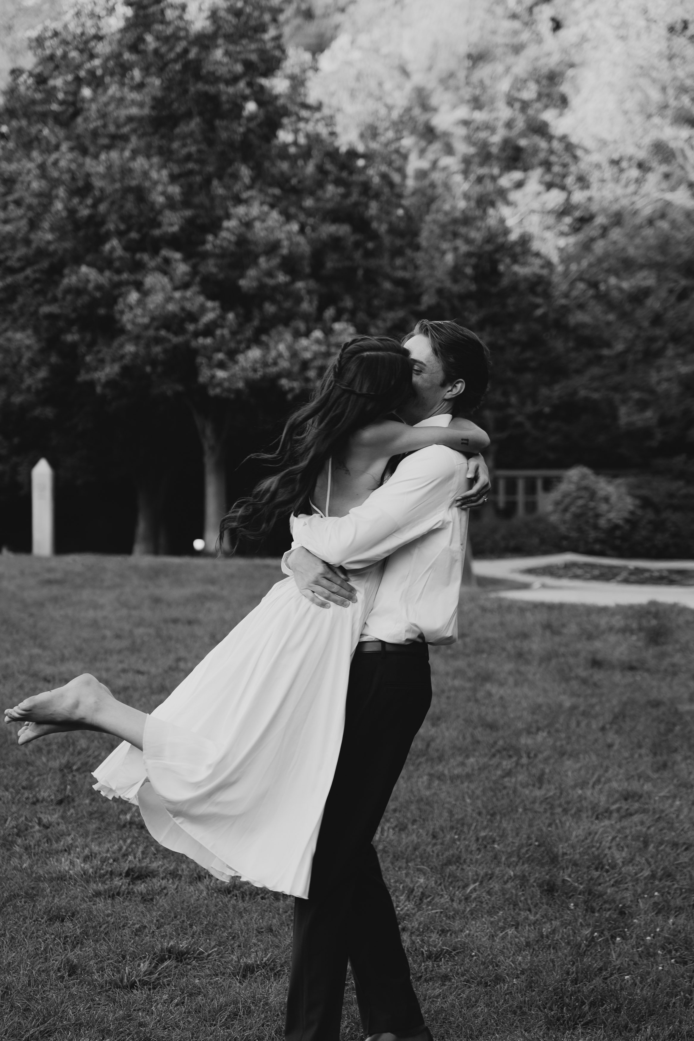 A man is holding a woman in a white dress in his arms, both are embracing and kissing outdoors in a park with trees in the background.