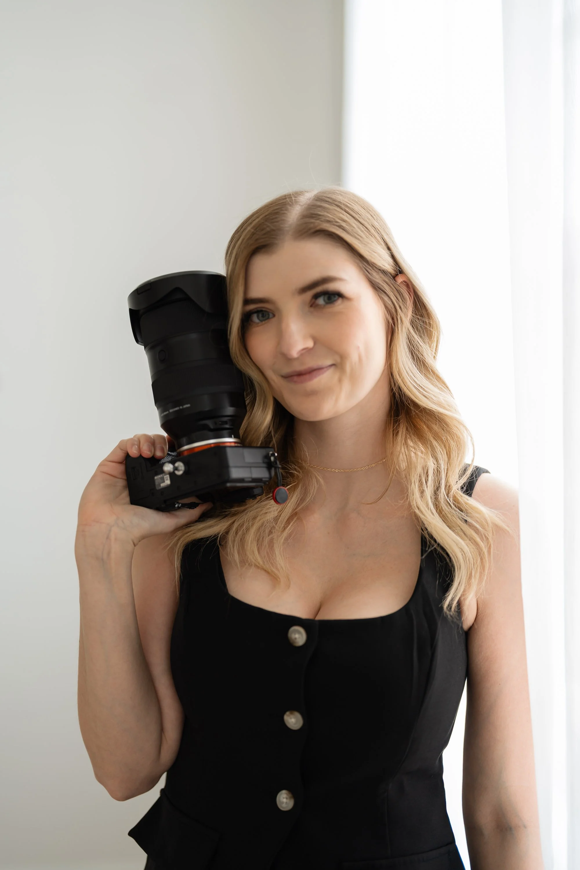 A young woman with blonde hair and blue eyes smiling while holding a professional camera with a large lens, standing in front of a neutral background with light coming from the right side.