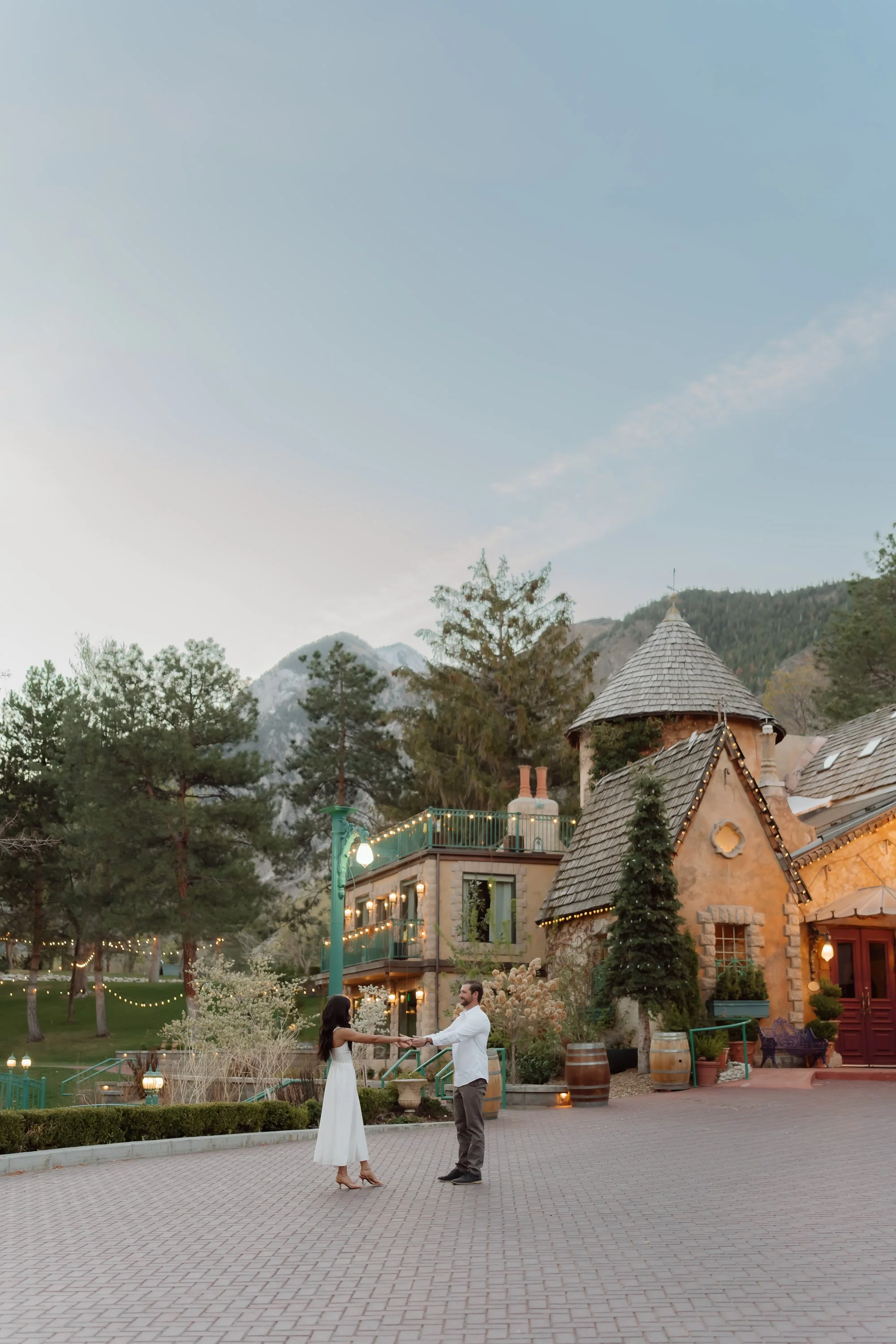 A couple dancing outdoors in front of a charming European-style building with lit windows and towers, surrounded by trees and mountain scenery, during dusk.