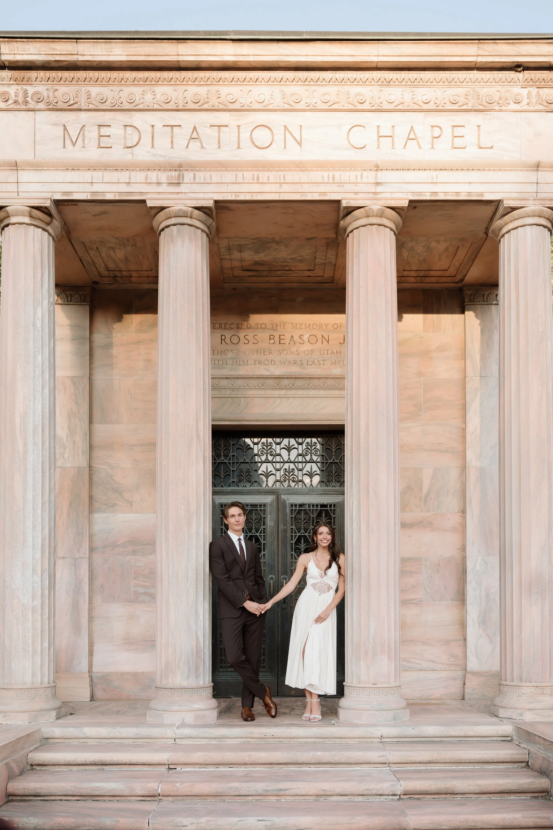A newlywed couple stands hand in hand in front of the Meditation Chapel with classical columns and ornate decoration, smiling and dressed in wedding attire.