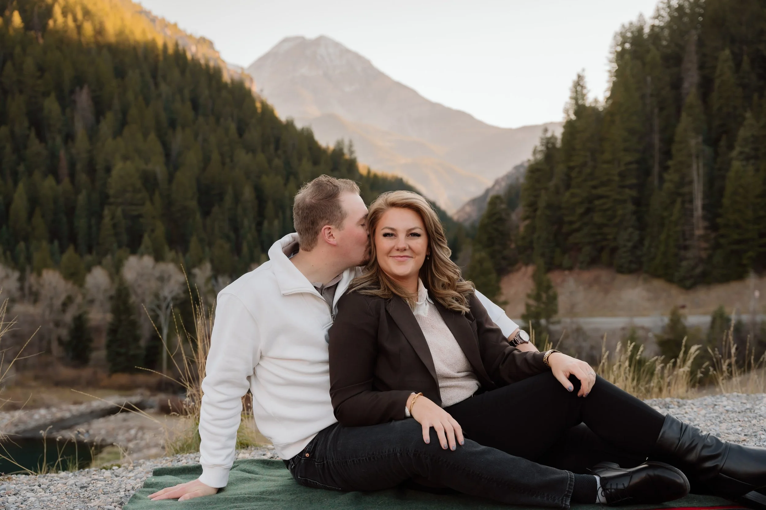 A couple sitting outdoors in a mountainous area with pine trees, one man kissing the woman on the cheek. The woman has long, wavy hair and is smiling at the camera. They are sitting on a blanket on rocky ground, with a mountain and trees in the backg