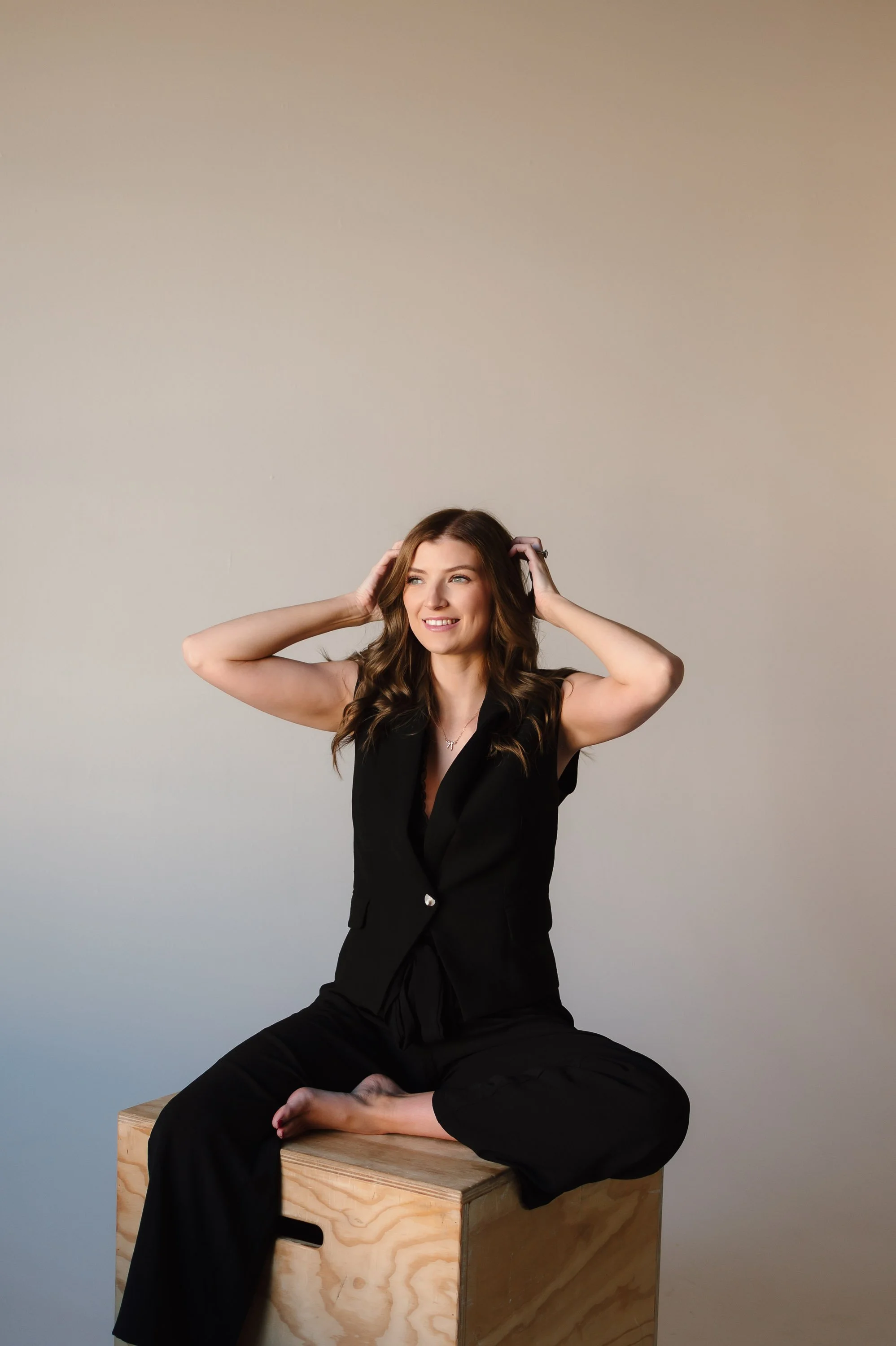 A woman sitting cross-legged on a wooden box, smiling, with her hands in her hair, wearing a black sleeveless outfit against a plain background.