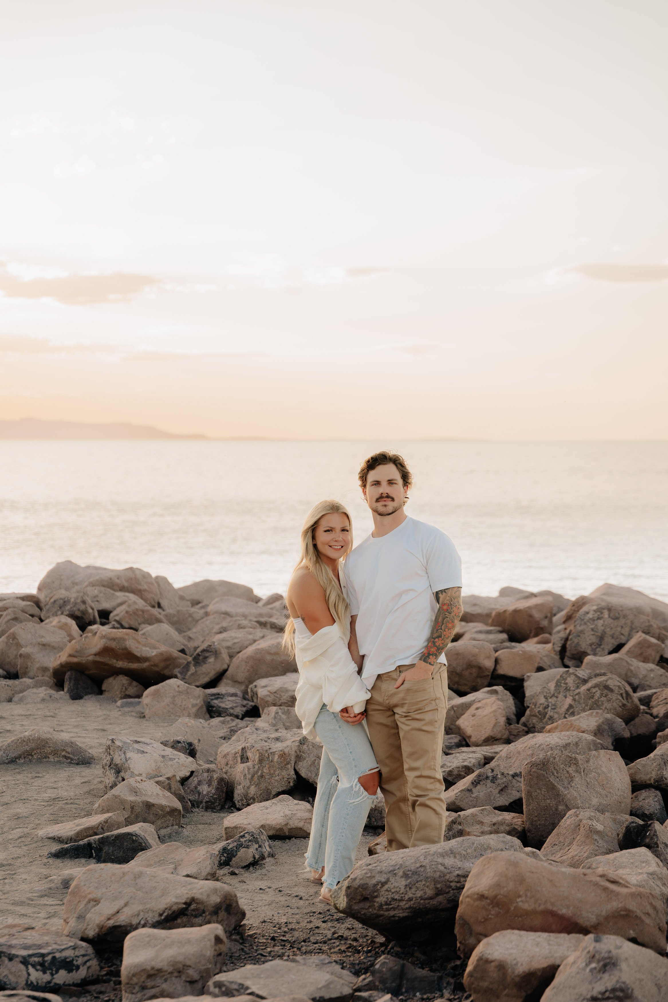 A young couple standing on a rocky beach at sunset, with the ocean and distant landforms in the background, smiling and embracing.