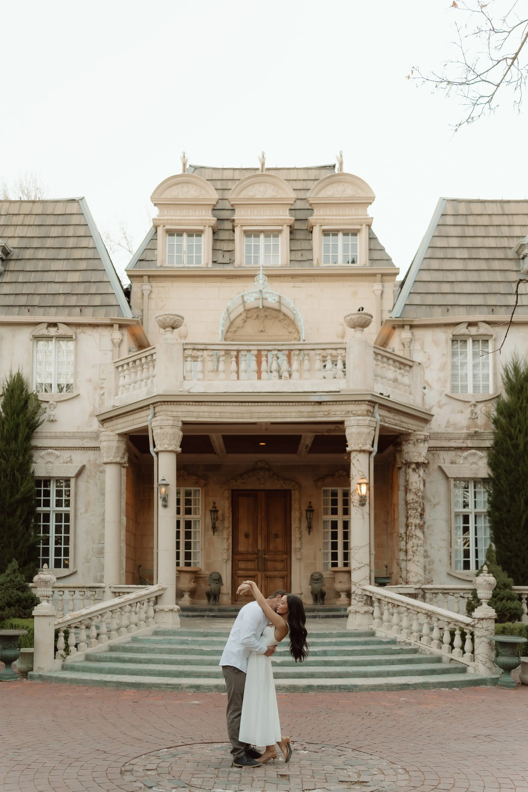 A couple dancing on the steps in front of an elegant, historic mansion with a stone facade, arched windows, and a balcony with balustrades. The mansion has a steeply pitched roof with dormer windows and decorative statues on the roofline. The couple 