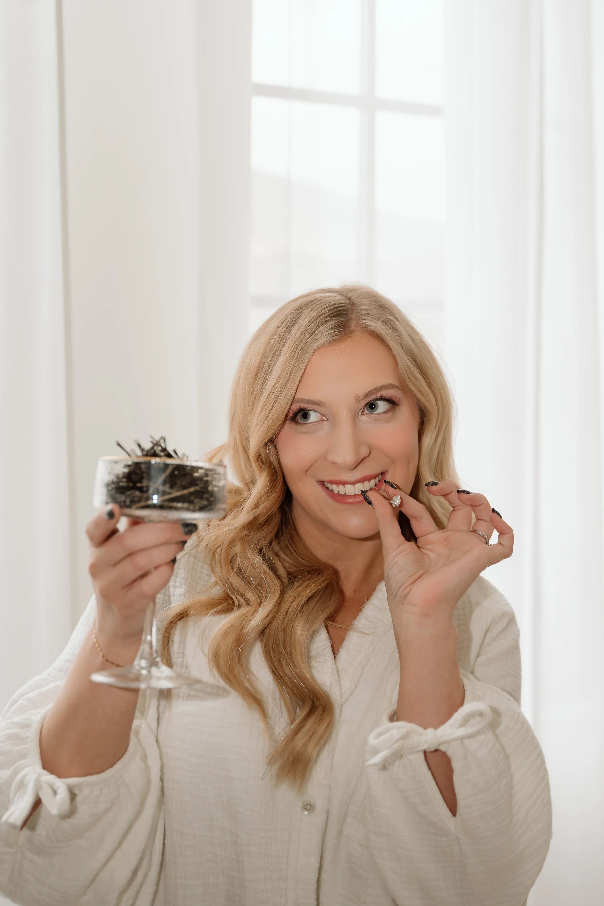 A woman with long blonde hair, smiling and applying lipstick, holding a glass container with makeup brushes, standing in front of windows with sheer curtains.