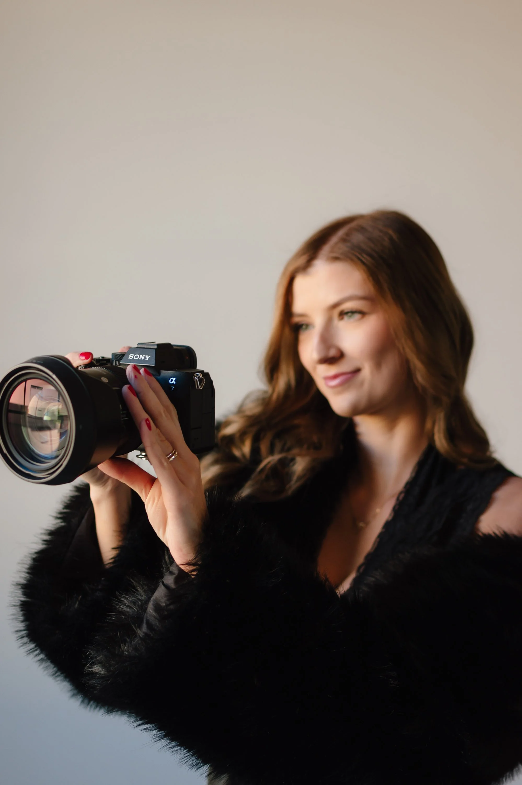 A woman with long brown hair looks at a Sony camera she is holding, wearing a black faux fur coat.