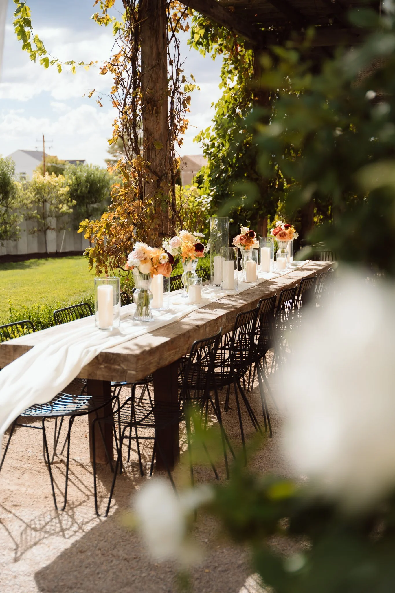 Outdoor dining table decorated with floral centerpieces and white candles, set in a garden with trees and lush greenery.