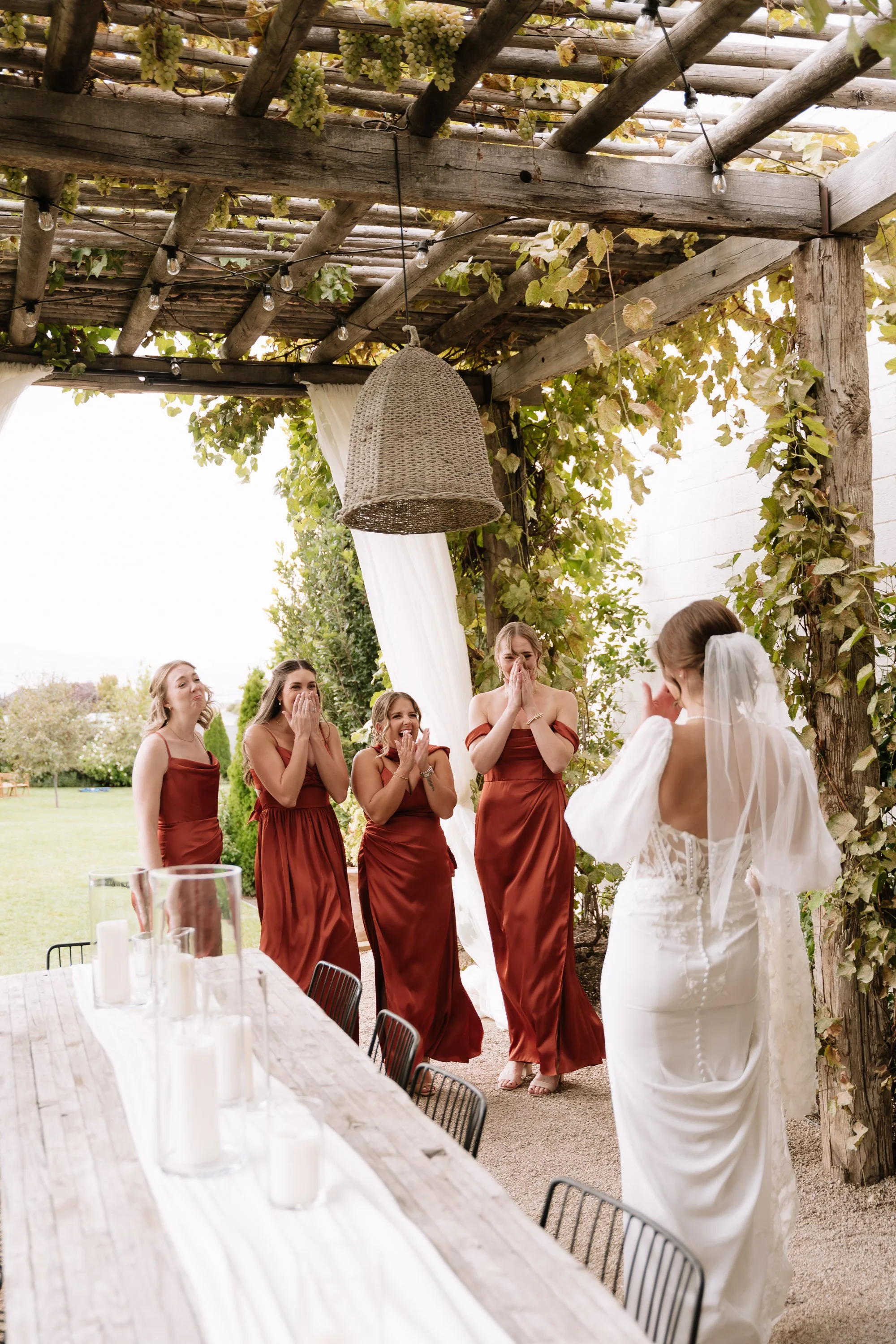 Bridesmaids and a bride at an outdoor wedding reception, with guests watching and reacting emotionally, under a wooden pergola decorated with string lights and greenery.