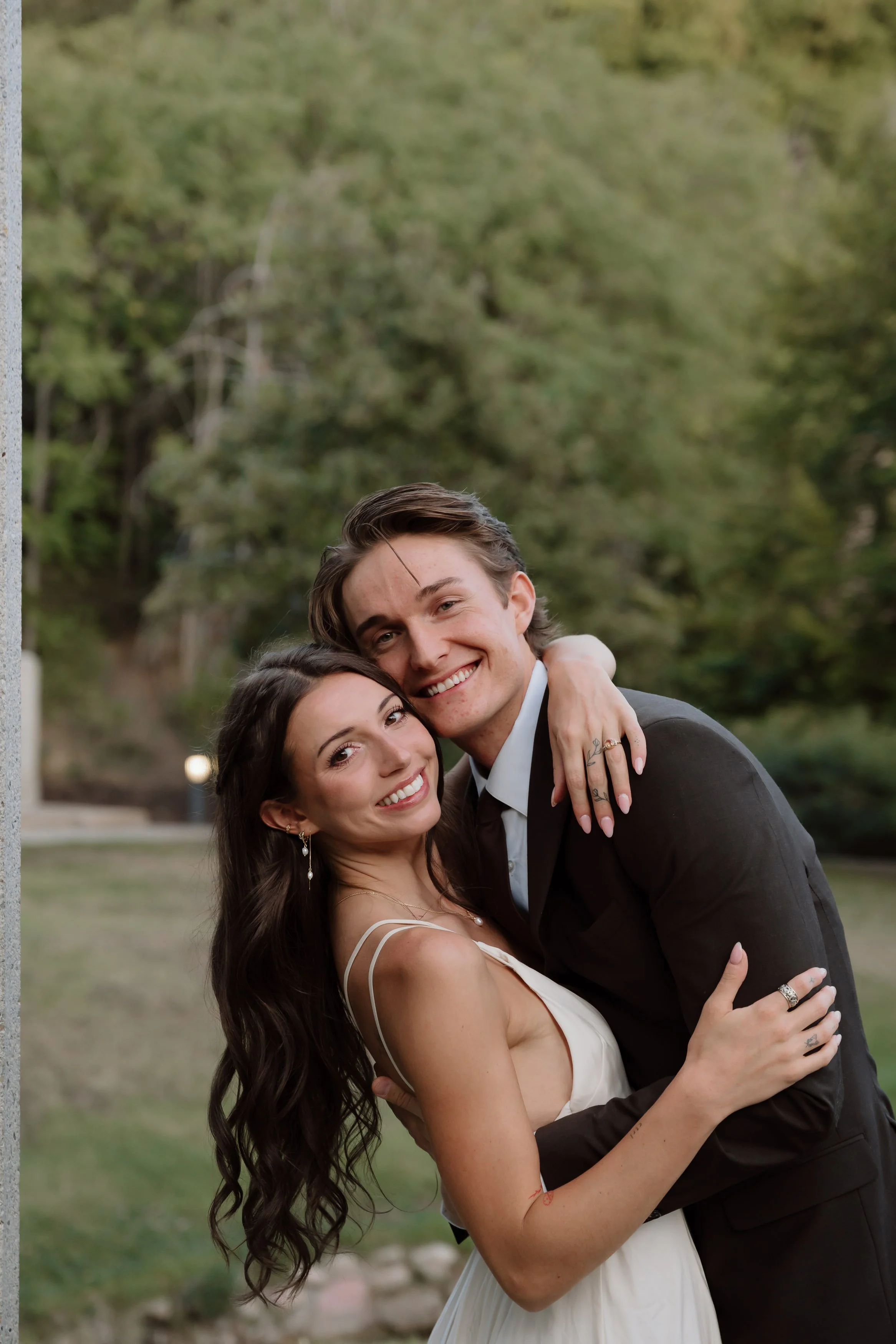 A happy couple is embracing outdoors, with trees in the background. The woman has long dark hair and is wearing a white dress with thin straps, and the man has short brown hair and is wearing a gray suit. They are smiling and looking at the camera.