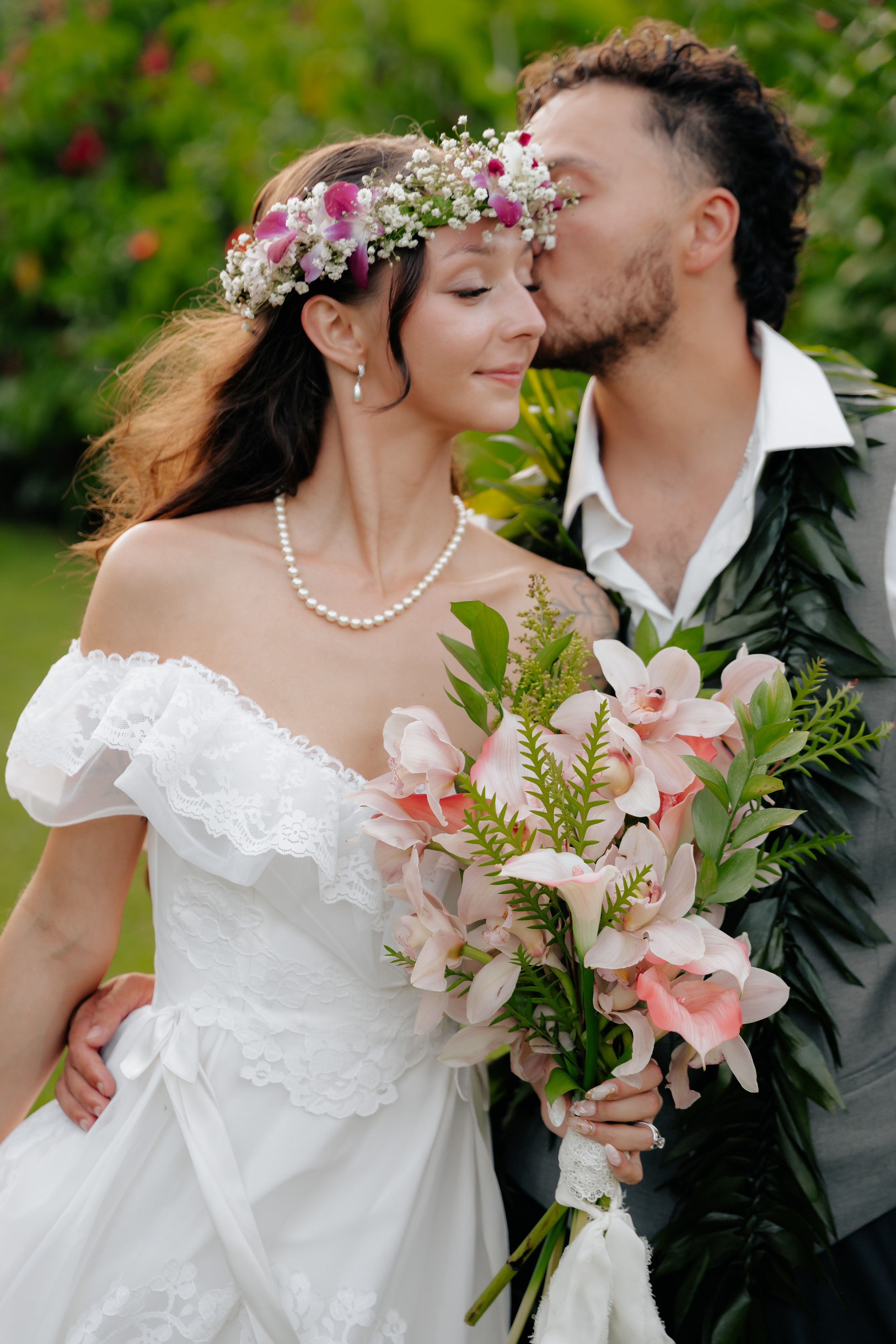 A bride and groom sharing an intimate moment, with the groom kissing the bride on the forehead. The bride wears an off-shoulder white wedding dress with lace details, a pearl necklace, and a floral crown. She holds a bouquet of pink and white calla lilies with greenery. The groom wears a white shirt and a dark vest, with green foliage draped over his shoulders. They are in a lush garden setting.