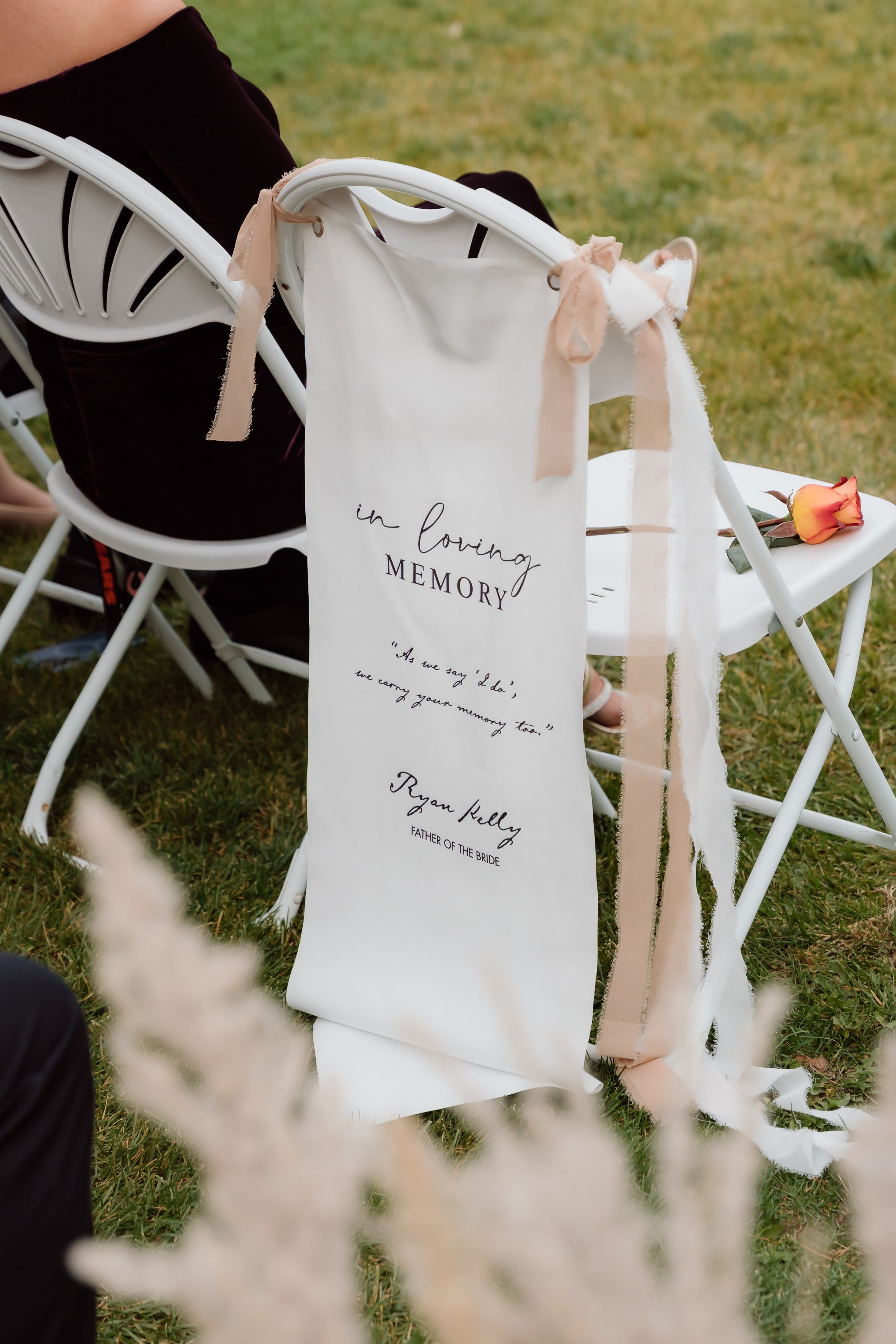 A white ribbon with black and cursive writing for a wedding memorial. A chair with beige and white ribbons is nearby, with a pink and orange rose on a white tablecloth. The scene is outdoors on grass.