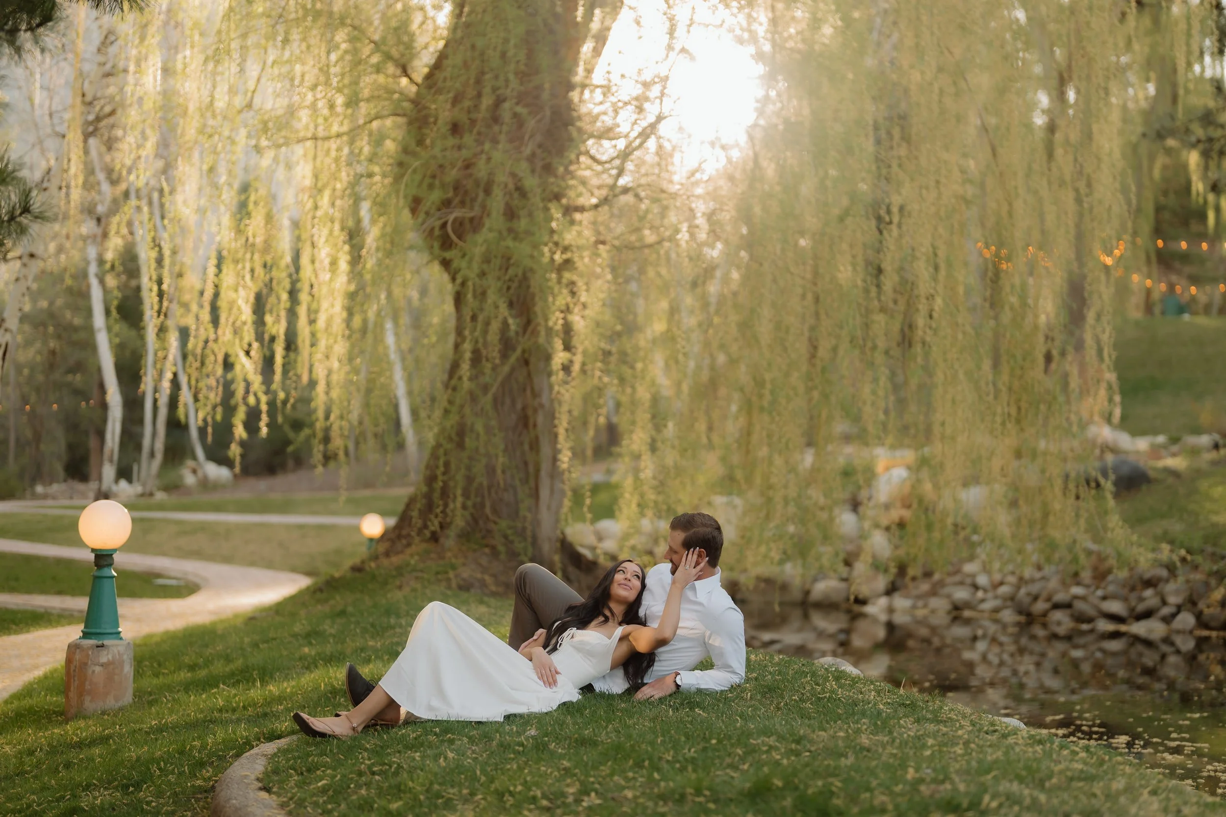 A couple sitting on grass near a pond, embracing each other, with trees and warm sunlight in the background.