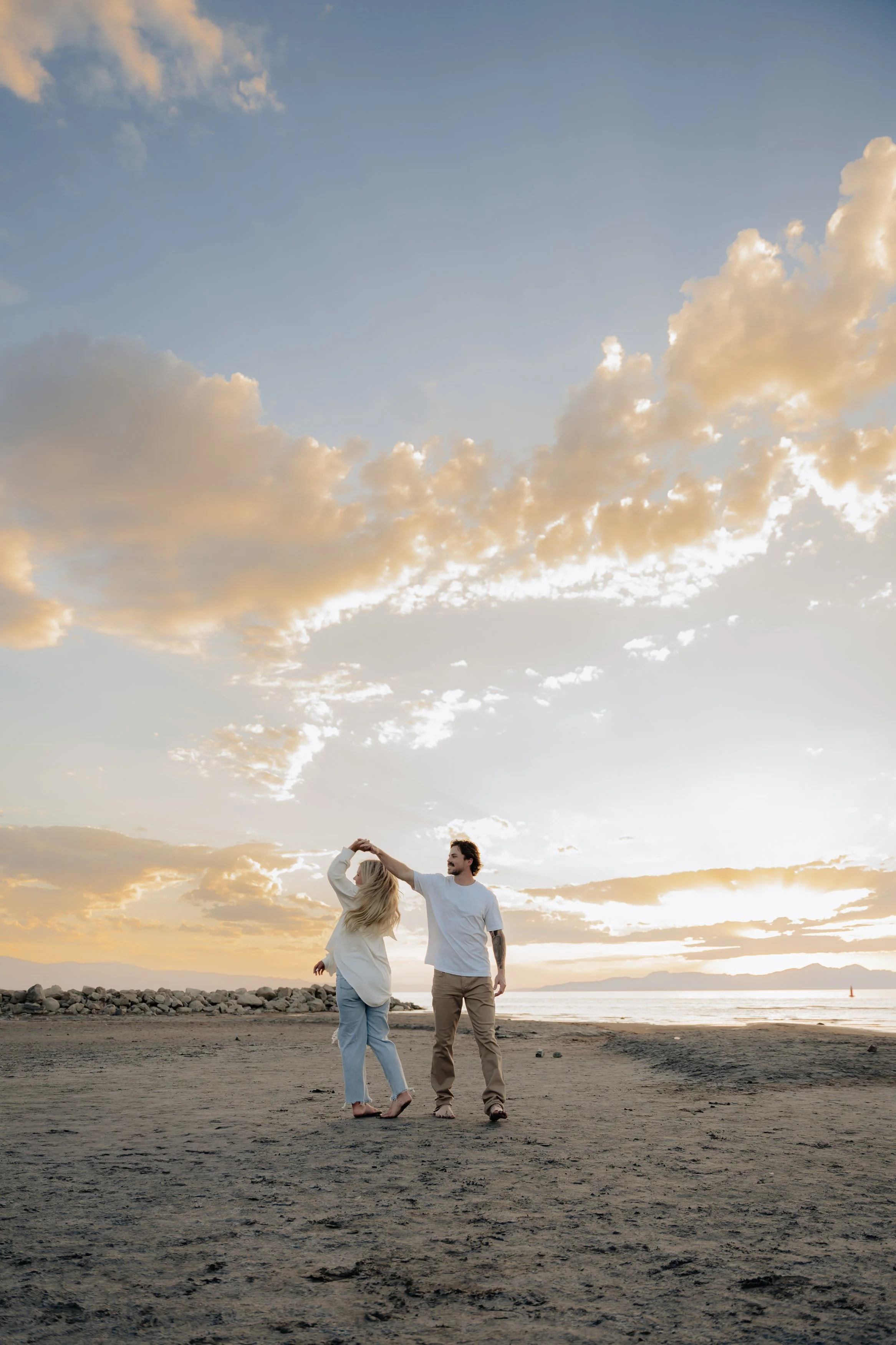 A couple walking on a sandy beach during sunset, with the man twirling the woman and both smiling. The sky features scattered clouds illuminated by the setting sun.