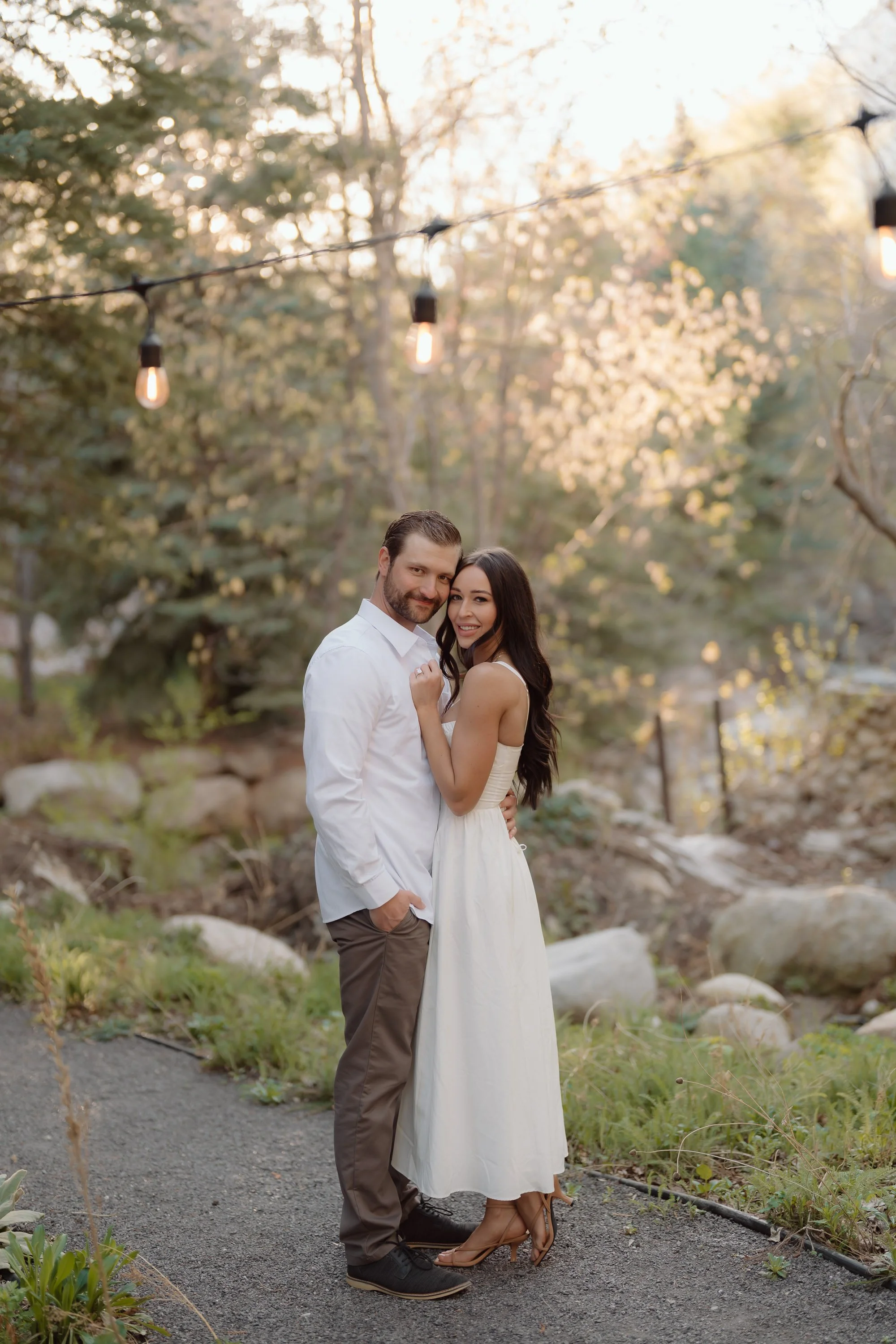 A couple standing outdoors among trees with string lights, smiling and posing together