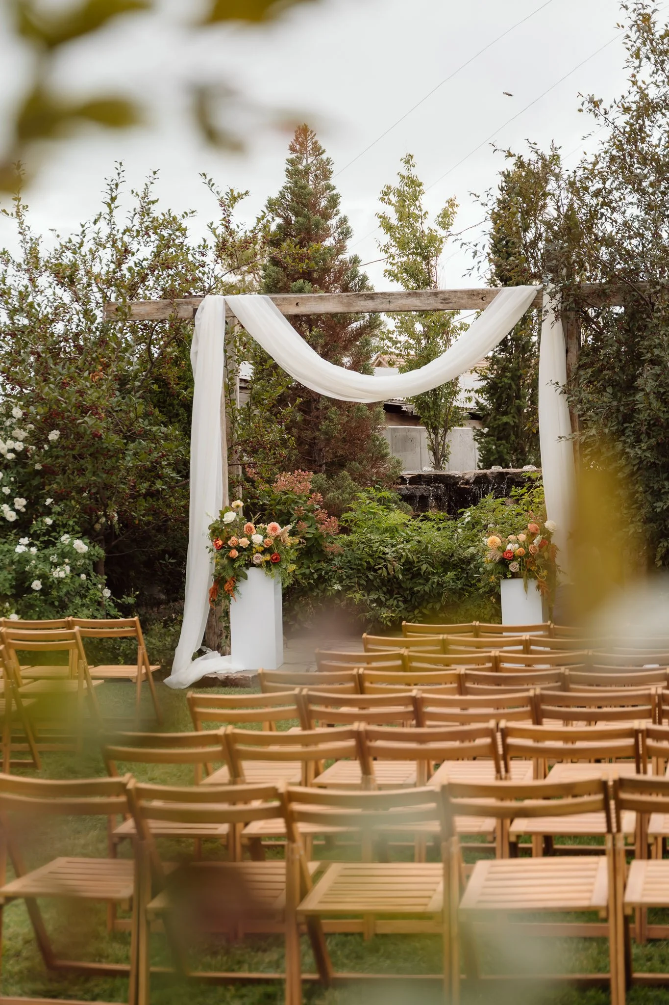 Outdoor wedding ceremony setup with wooden chairs arranged in rows, a decorated arch with white fabric and flowers, and lush green trees in a garden.