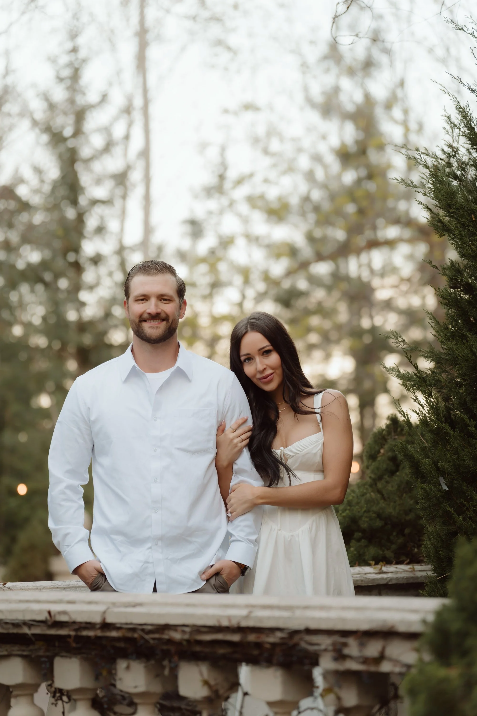 A young man and woman standing close together outdoors in a park or garden with trees in the background. The man has light brown hair, a beard, and is wearing a white shirt. The woman has long dark hair and is wearing a light-colored dress. They are 